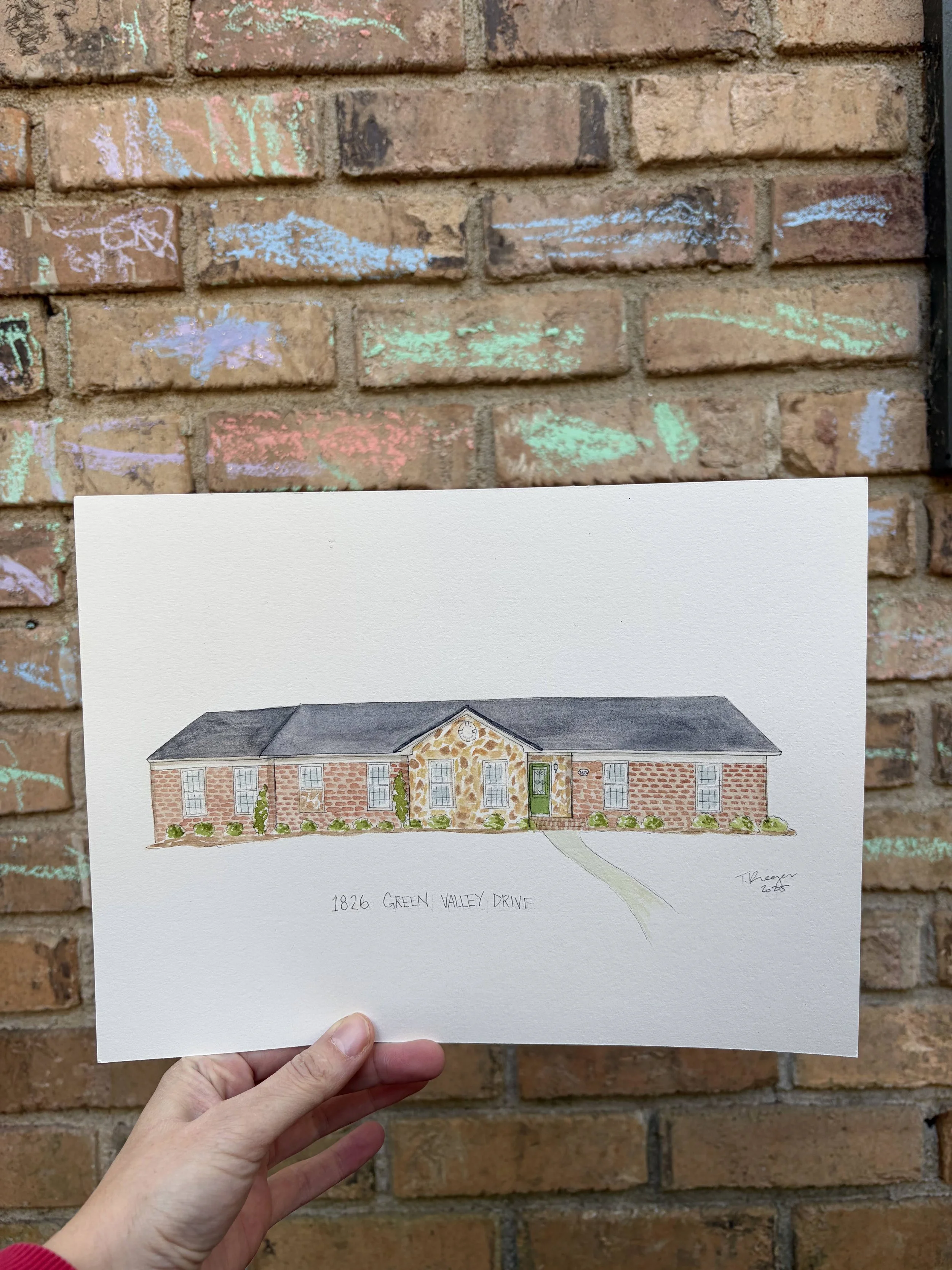 A person holding a colored pencil drawing of a house with the address 1826 Green Valley Drive in front of a brick wall with colorful chalk drawings.