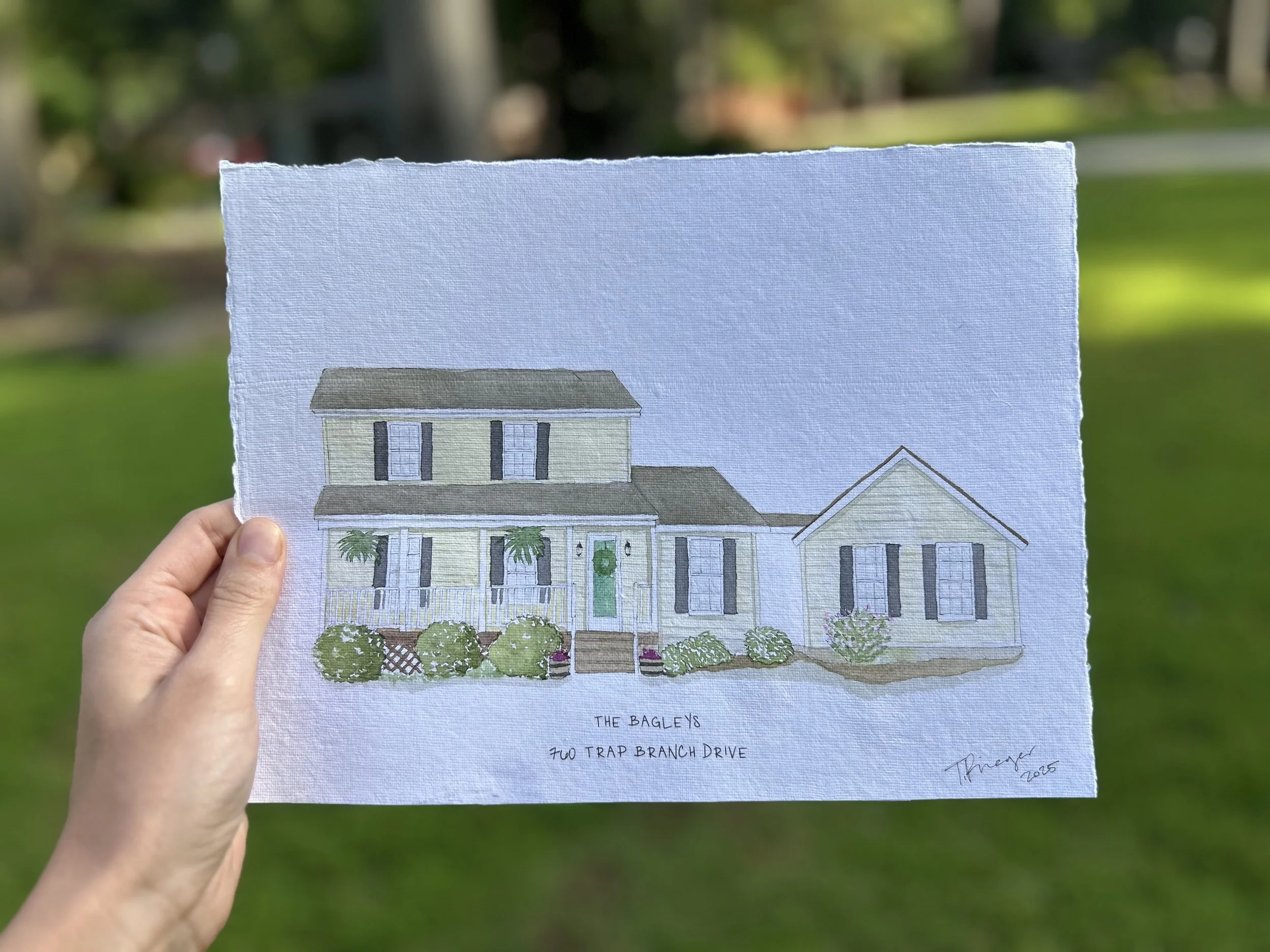 A hand holding a paper with a colored architectural drawing of a two-story house with a porch, front steps, and plants in front, against a background of green grass and trees.