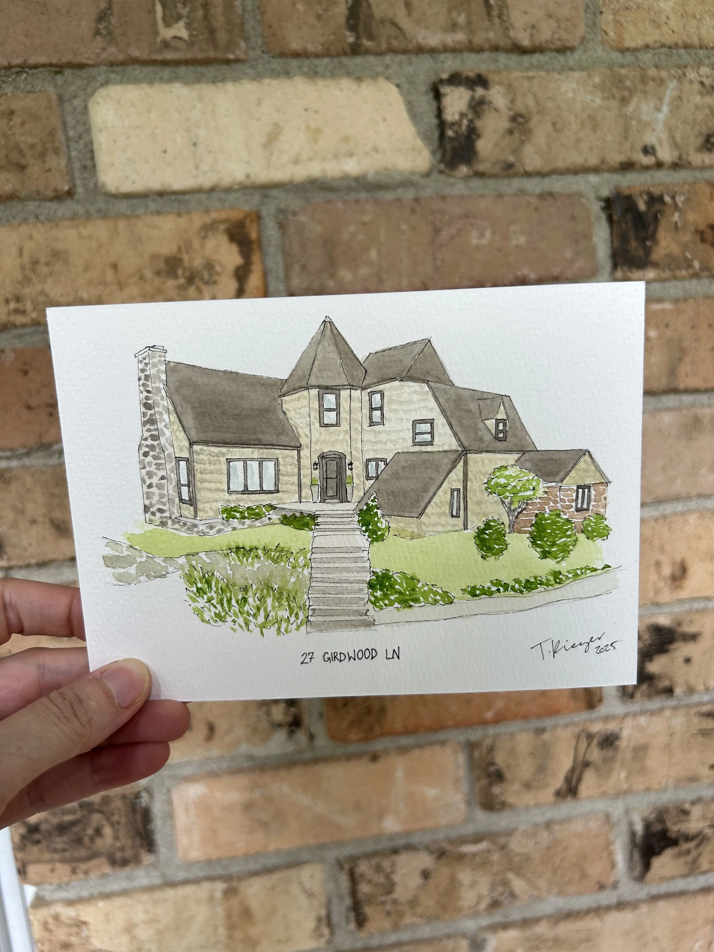 A hand holding a watercolor illustration of a house with a brick chimney and front yard, set against a brick wall background.