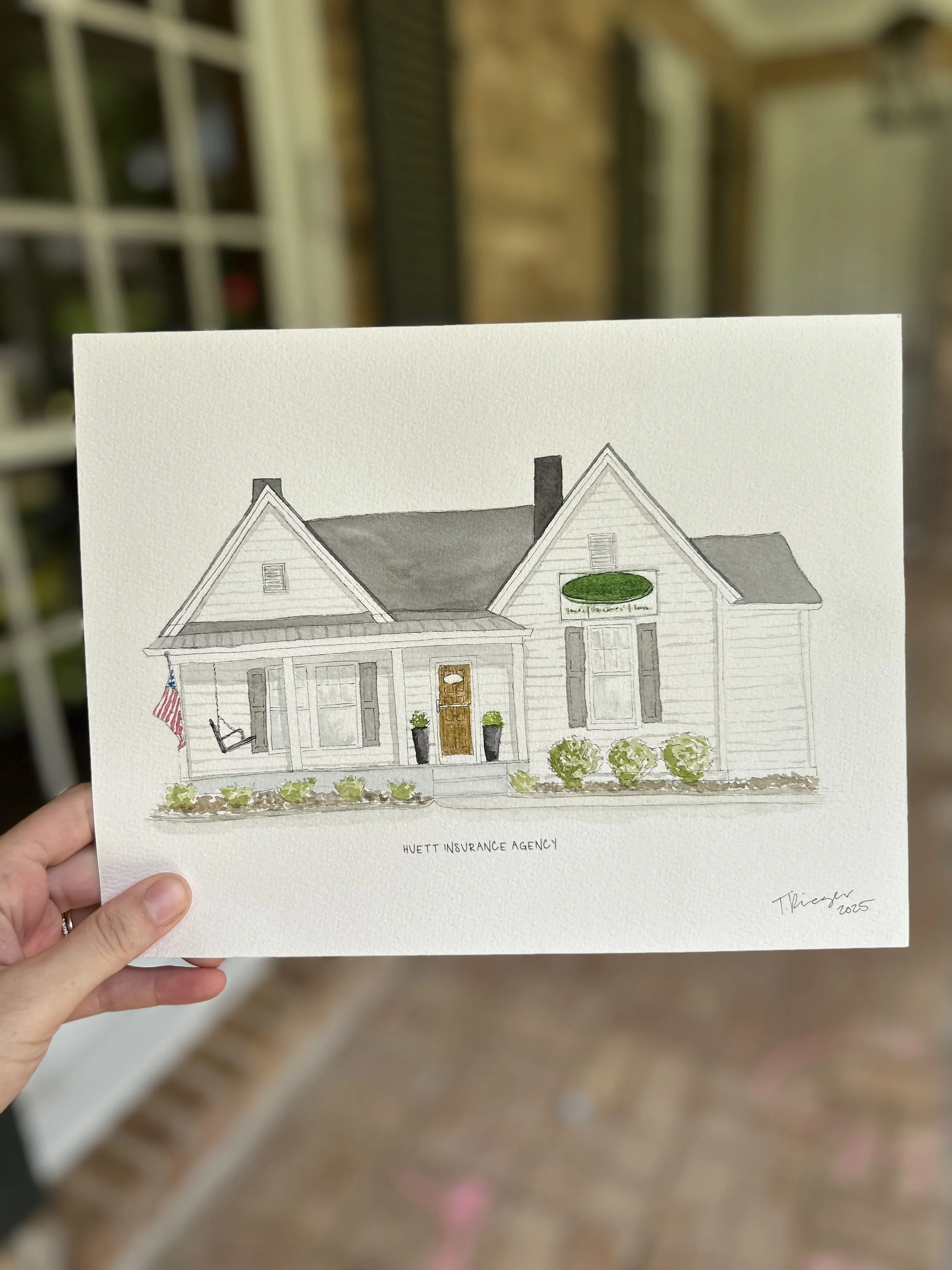 A hand holding a watercolor illustration of a white house with gray roof and front porch, with American flag, two black planters, and green bushes. The illustration is signed "T. Hiegg 2023" at the bottom right.