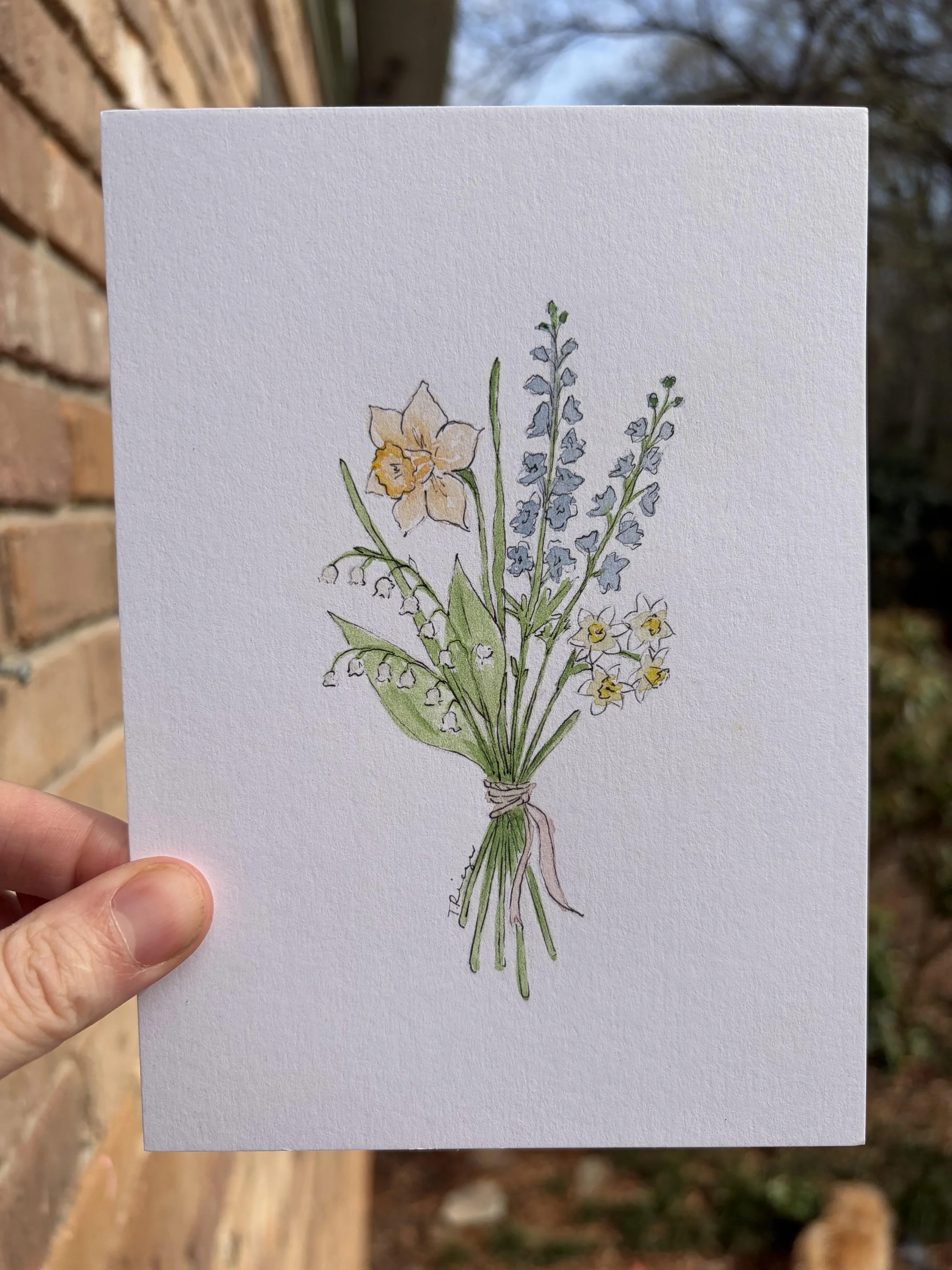 Hand holding a watercolor illustration of a bouquet of spring flowers on white paper, with a brick wall and trees in the background.