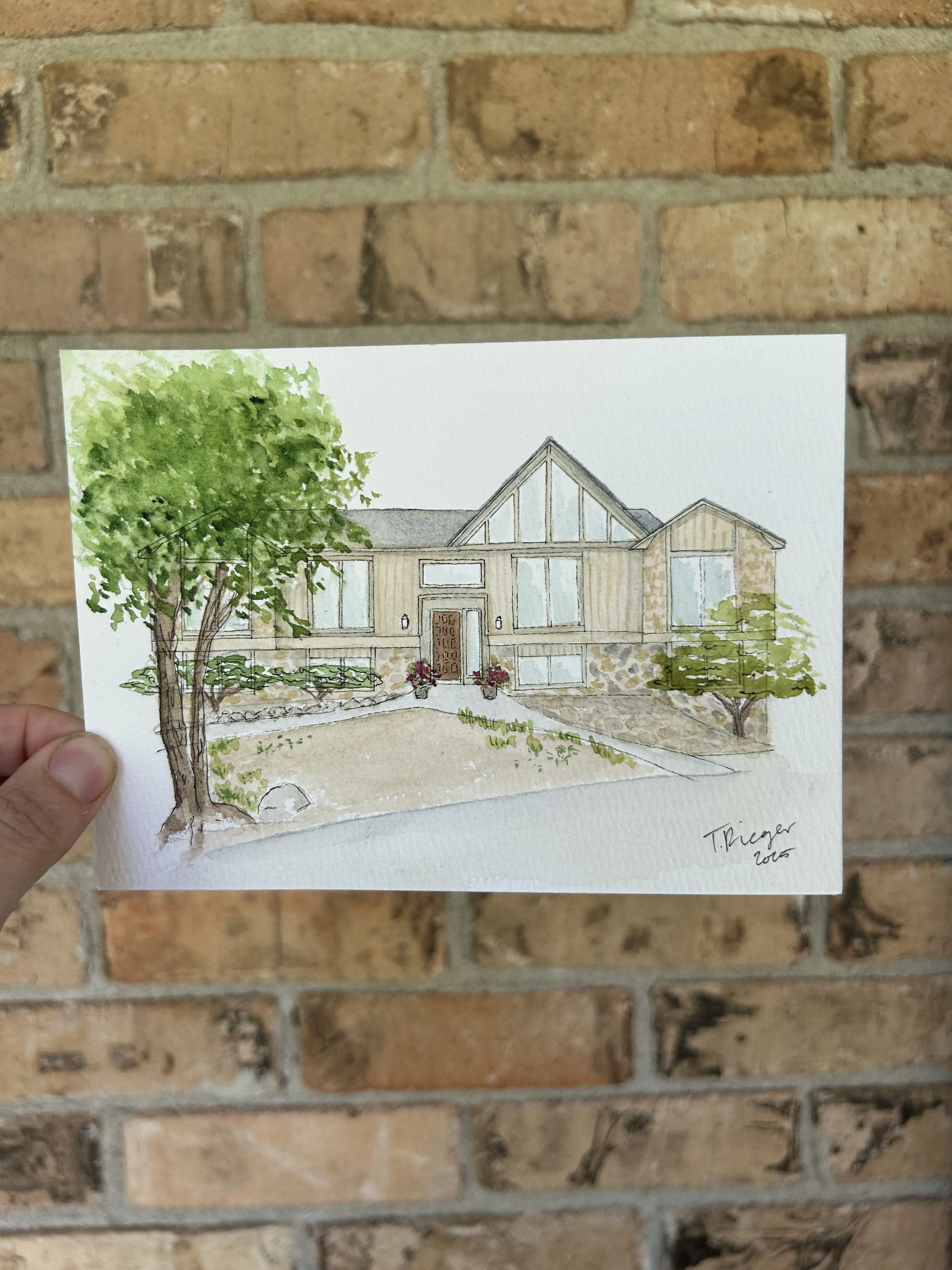 Colorful watercolor illustration of a modern house with large glass windows, a peaked roof, and a front door, with trees and shrubs in the yard, signed by T. Priger in 2015, held against a brick wall.