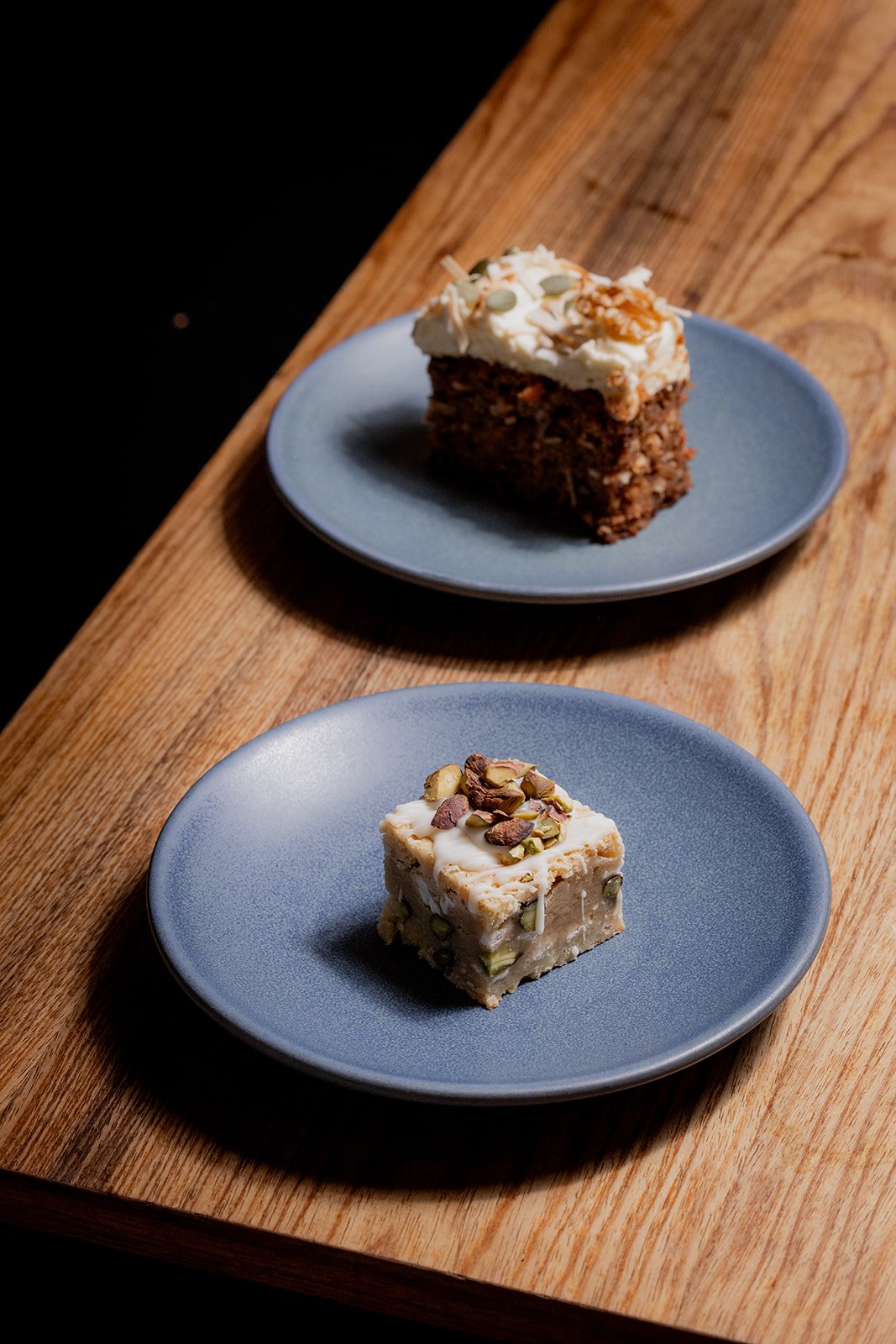 Two dessert plates on a wooden table, one with a slice of carrot cake topped with cream and nuts, and another with a pistachio-topped dessert square.
