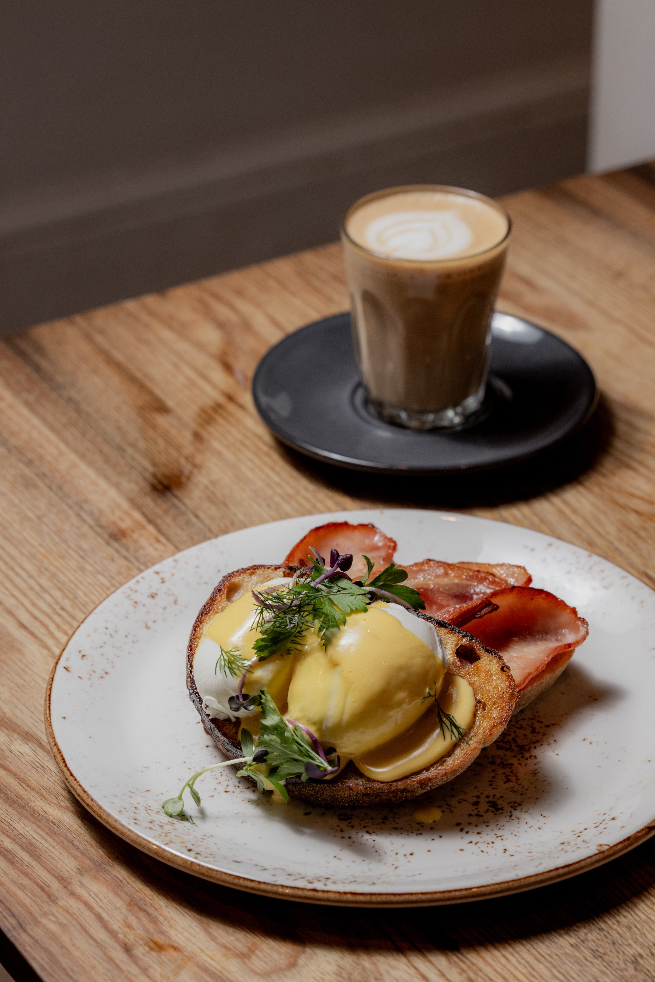 Eggs Benedict with hollandaise sauce, served with slices of ham on toast, garnished with greens, and a latte in a glass on a black saucer on a wooden table.