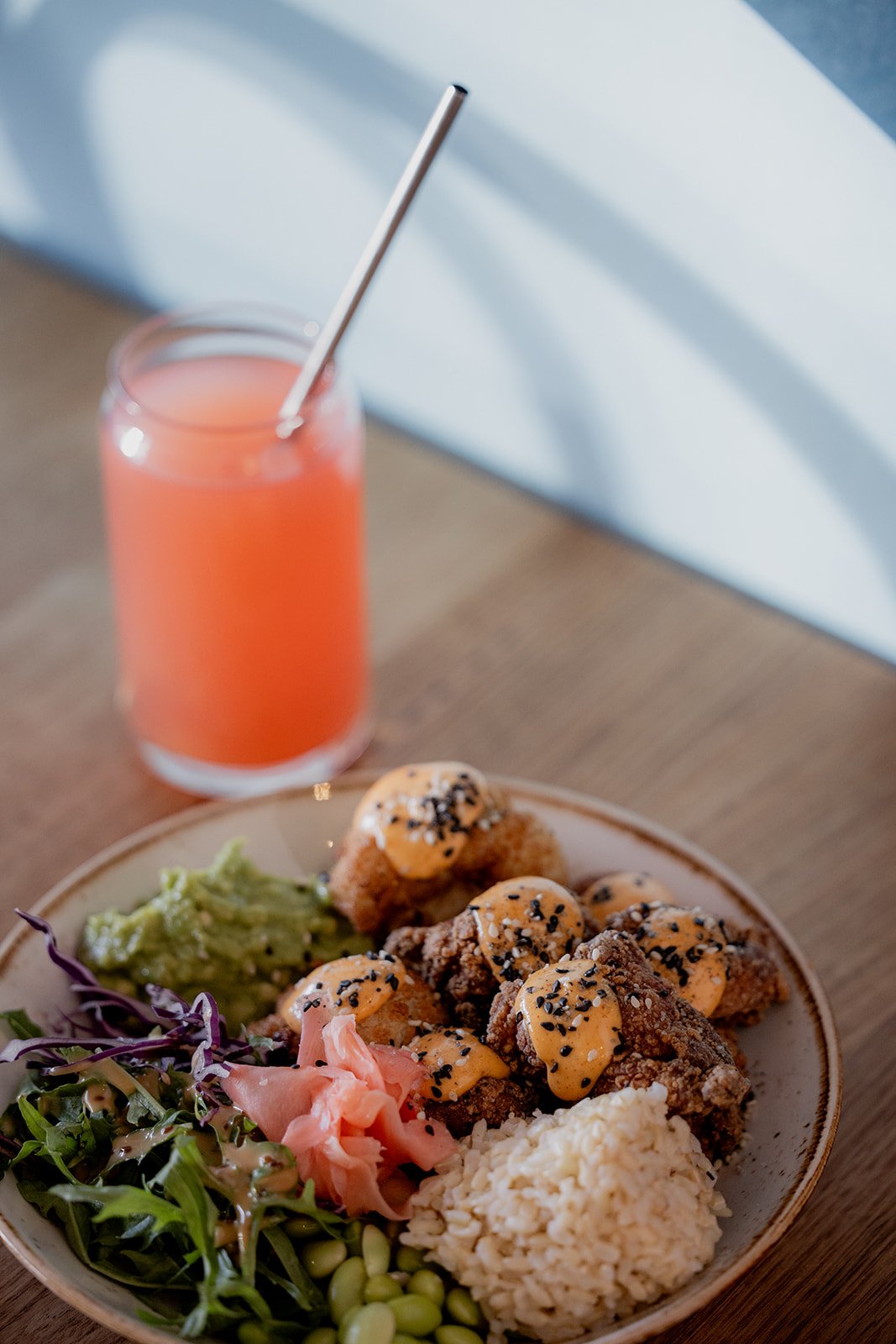 A plate with fried chicken topped with creamy sauce and sesame seeds, served with rice, guacamole, pickled ginger, and greens. A glass of pink beverage with a metal straw is in the background.