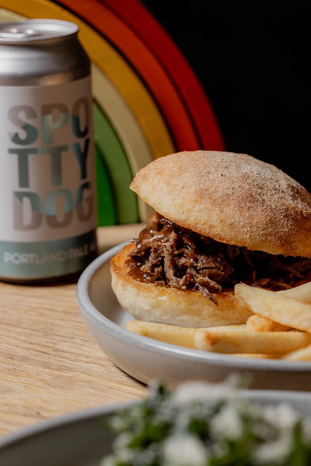 Pulled beef sandwich on a white plate with fries, a can labeled "Spotty Dog Portland Pale Ale," and a colorful striped background.