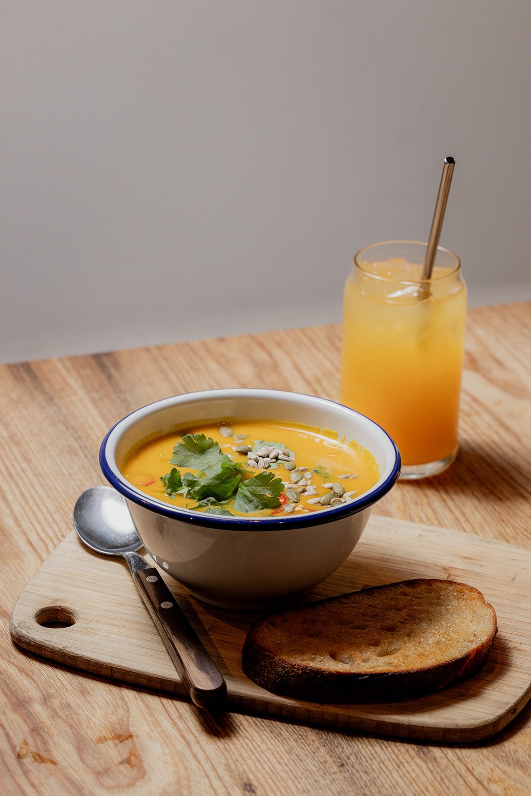 A bowl of orange soup garnished with seeds and herbs, placed on a wooden board with a spoon and a slice of bread. A glass of orange juice with a metal straw is in the background.