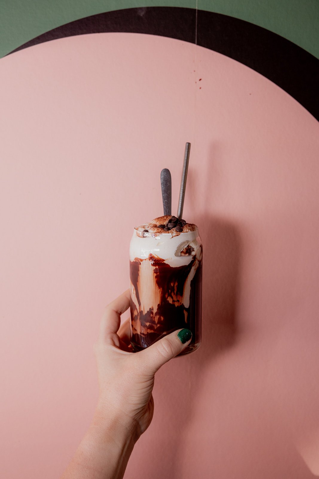 Hand holding a glass of iced chocolate drink with whipped cream and chocolate syrup against a pink background.