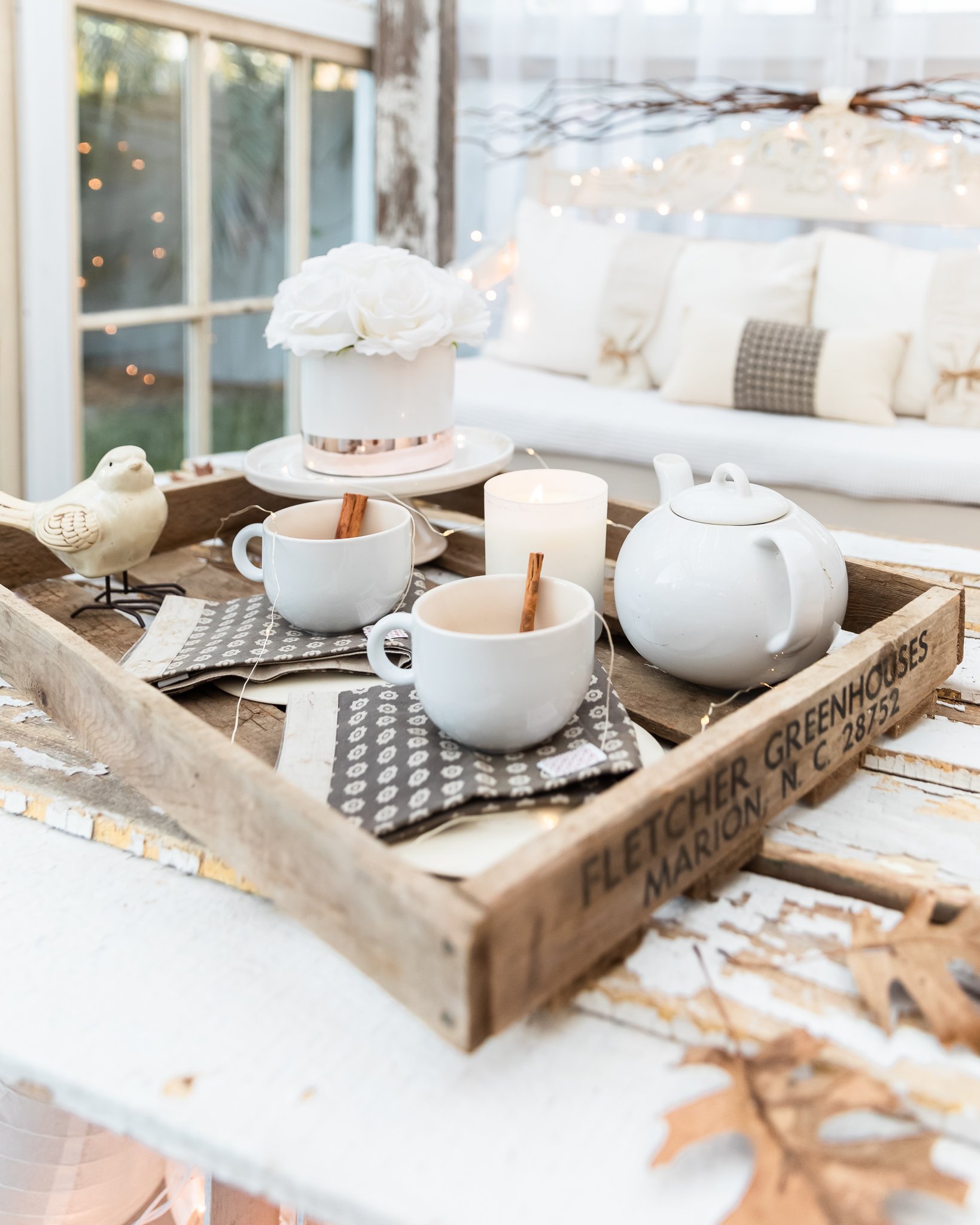 Decorative breakfast tray with white teapot, two white cups with cinnamon sticks, a white candle, a flower arrangement, a ceramic bird, and a napkin. In the background, a cozy seating area with cushions and string lights is visible.