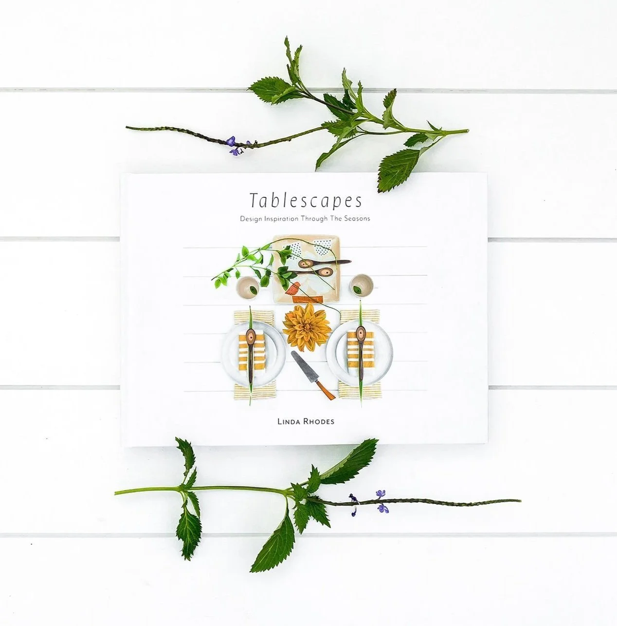Book titled "Tablescapes: Design Inspiration Through The Seasons" by Linda Rhodes, placed on a white surface with green leaves and purple flowers around it.