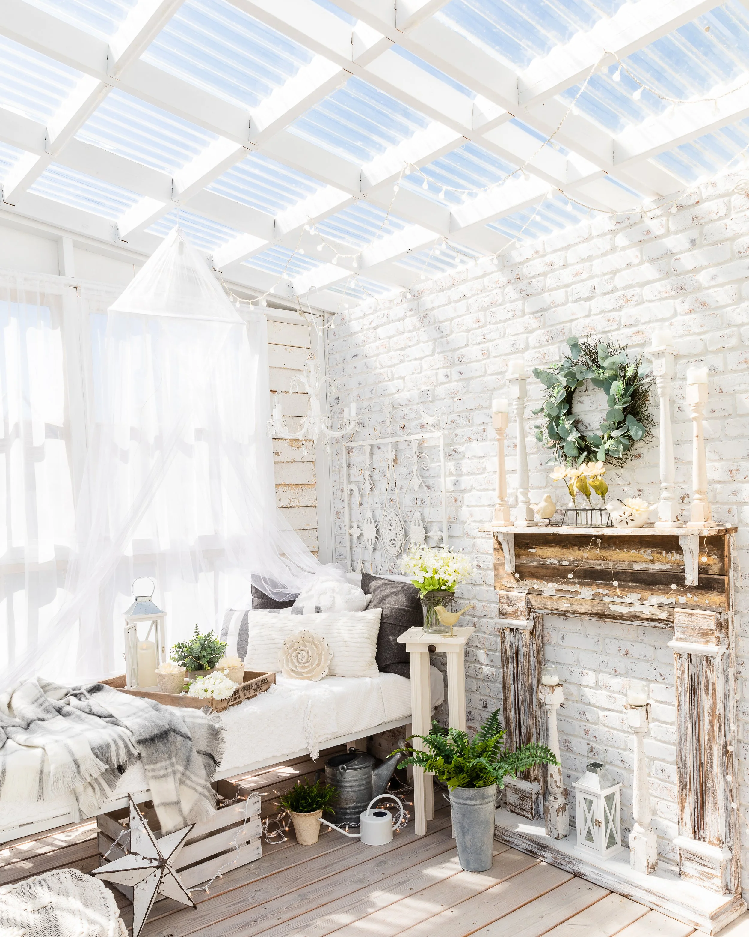 Bright sunroom with a white brick wall, featuring a cozy daybed with pillows and a blanket. Decor includes hanging sheer curtains, rustic wooden fireplace, greenery, lanterns, and potted plants.