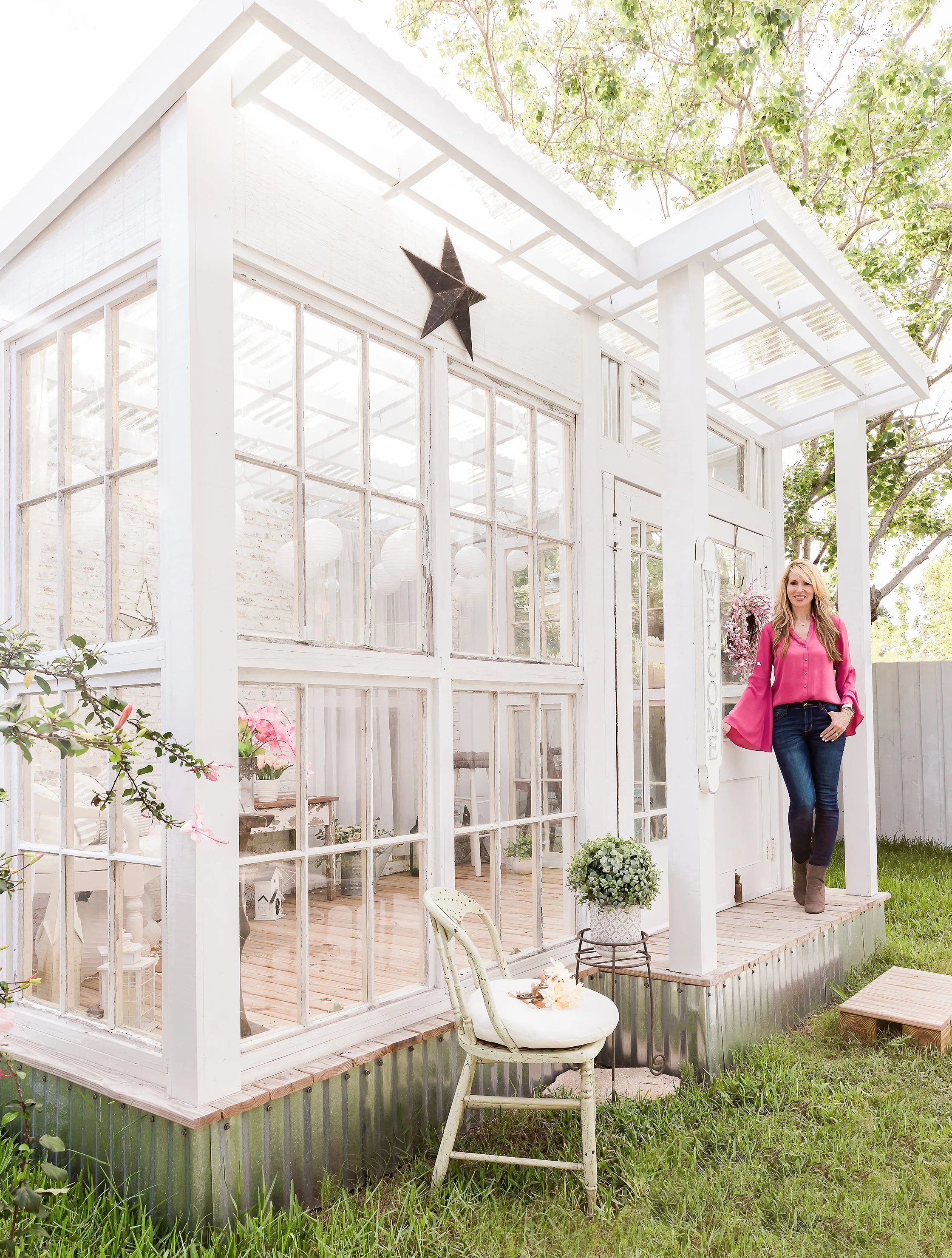 Stylish woman in pink standing outside a greenhouse with large windows, a black star decoration, and a rustic chair with a cushion on the grass.
