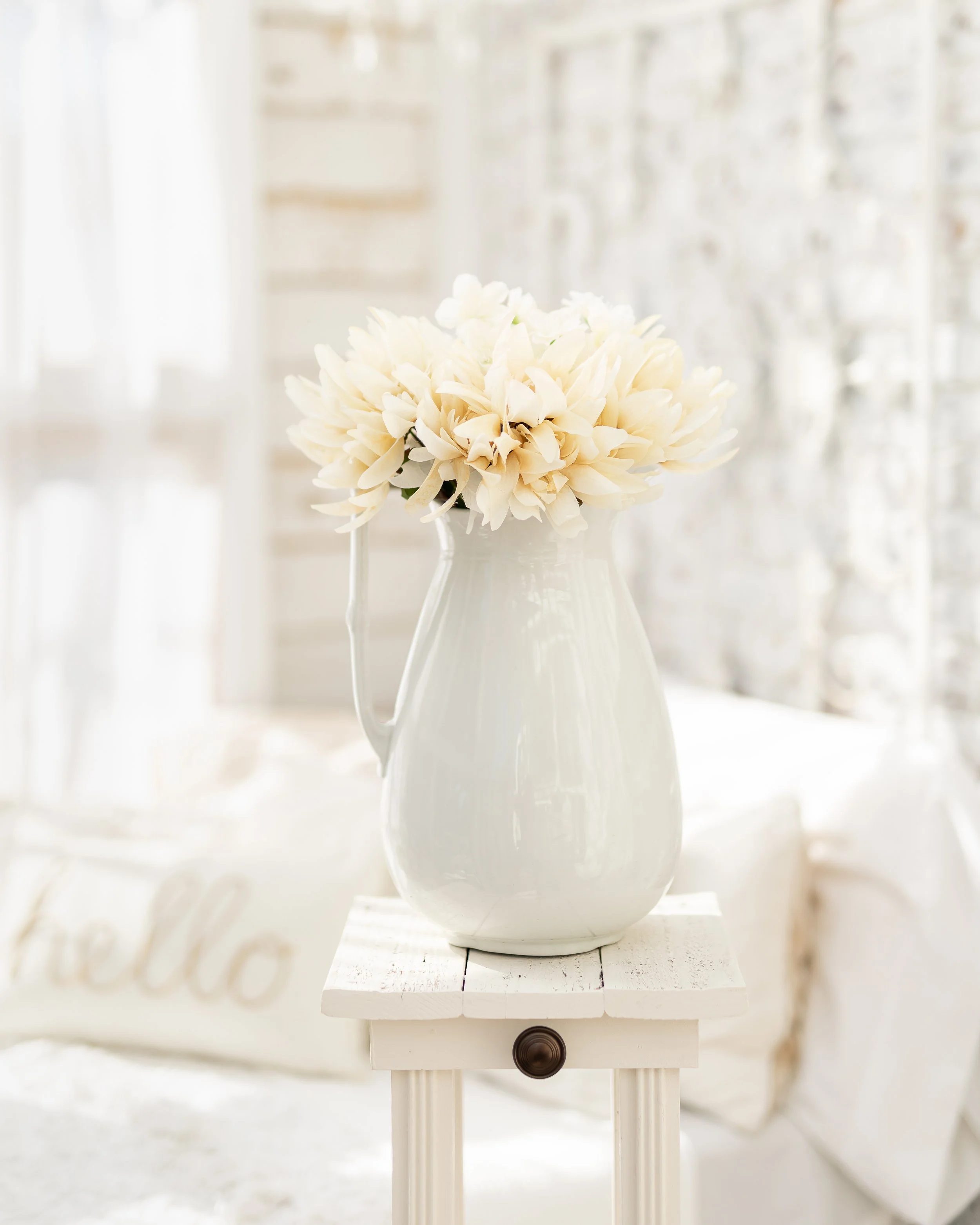 Cream-colored flowers in an antique white ceramic pitcher on a small repurposed table with a soft, light-filled background featuring a pillow with the word 'hello'.