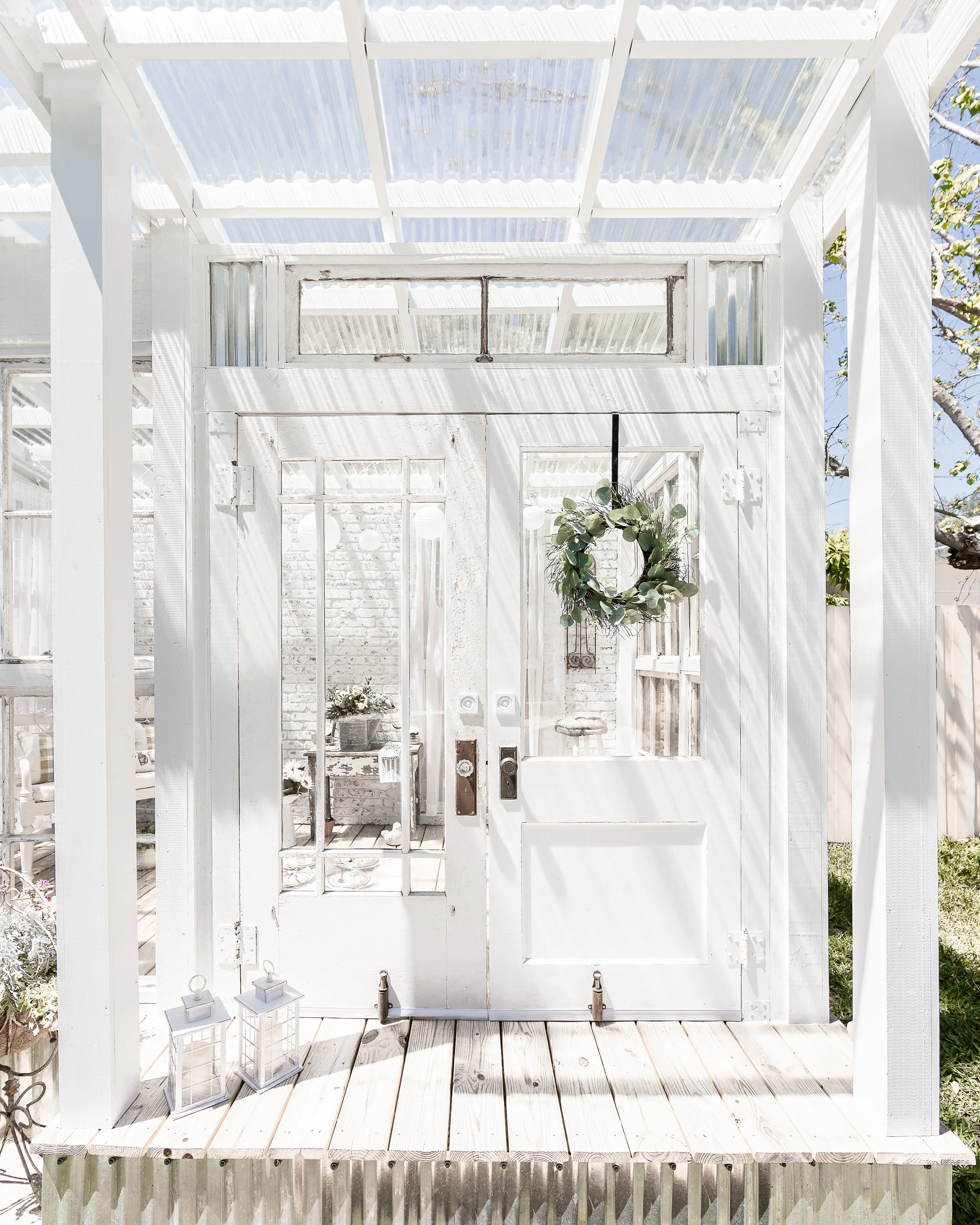 White wooden greenhouse with glass doors and a wreath hanging on the door, decorated with greenery, and two lanterns on the wooden deck outside.