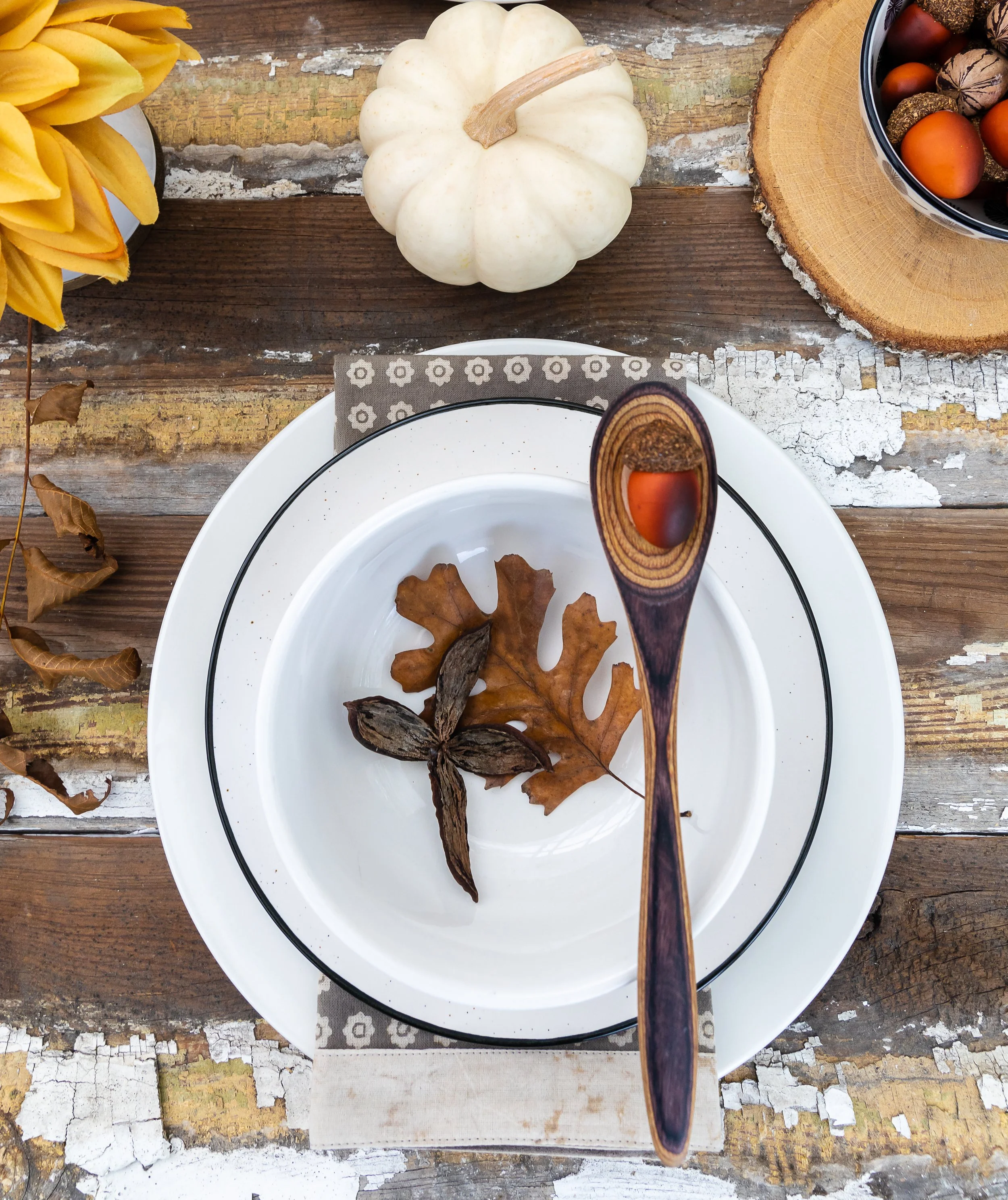 An autumn-themed table setting with a white pumpkin, dried leaves, dried seed pods, a bowl of acorns, and a plate with a wooden spoon, all on a rustic wooden table.
