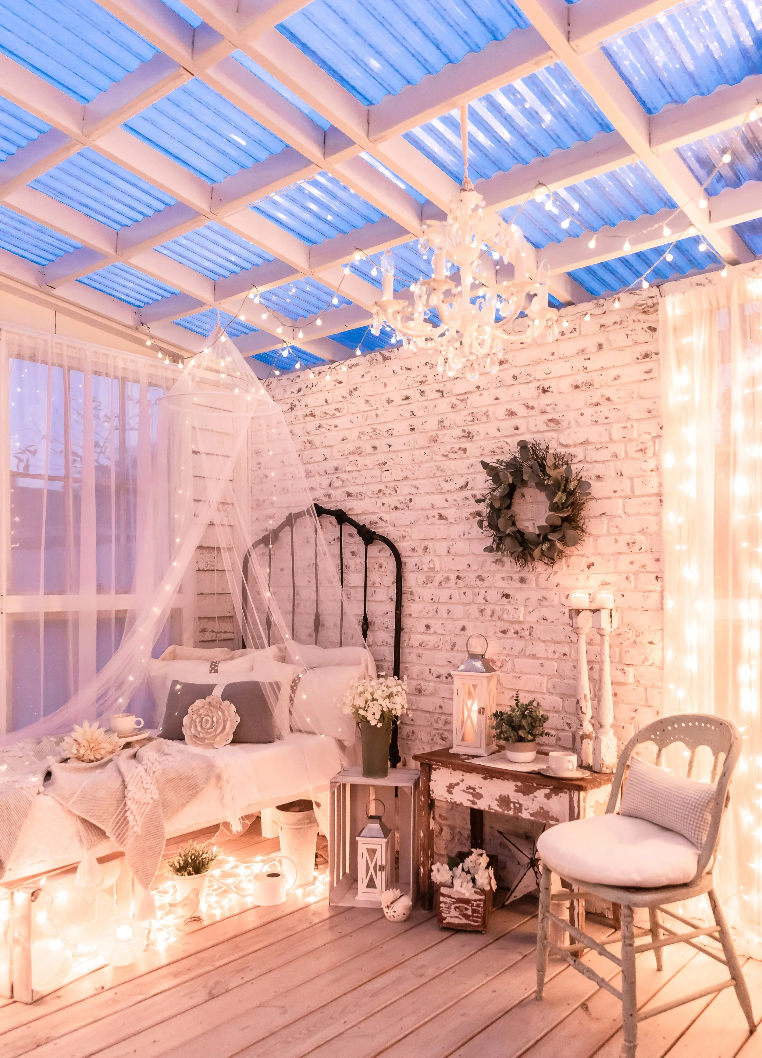 A cozy, decorated indoor space with a white brick wall, a white daybed with soft pillows and sheer fabric drapes, a small vintage wooden table with candles and plants, string lights, a chandelier hanging from the ceiling, and fairy lights along the r