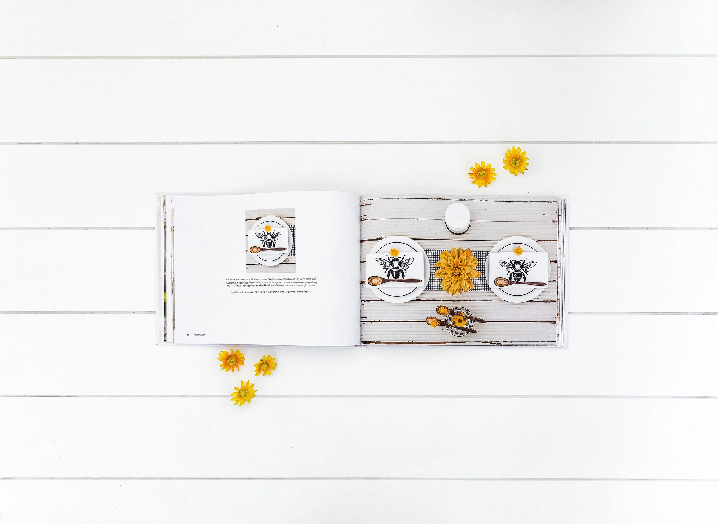 Open book on a white wooden table with a table setting featuring bee-themed plates and yellow flowers.