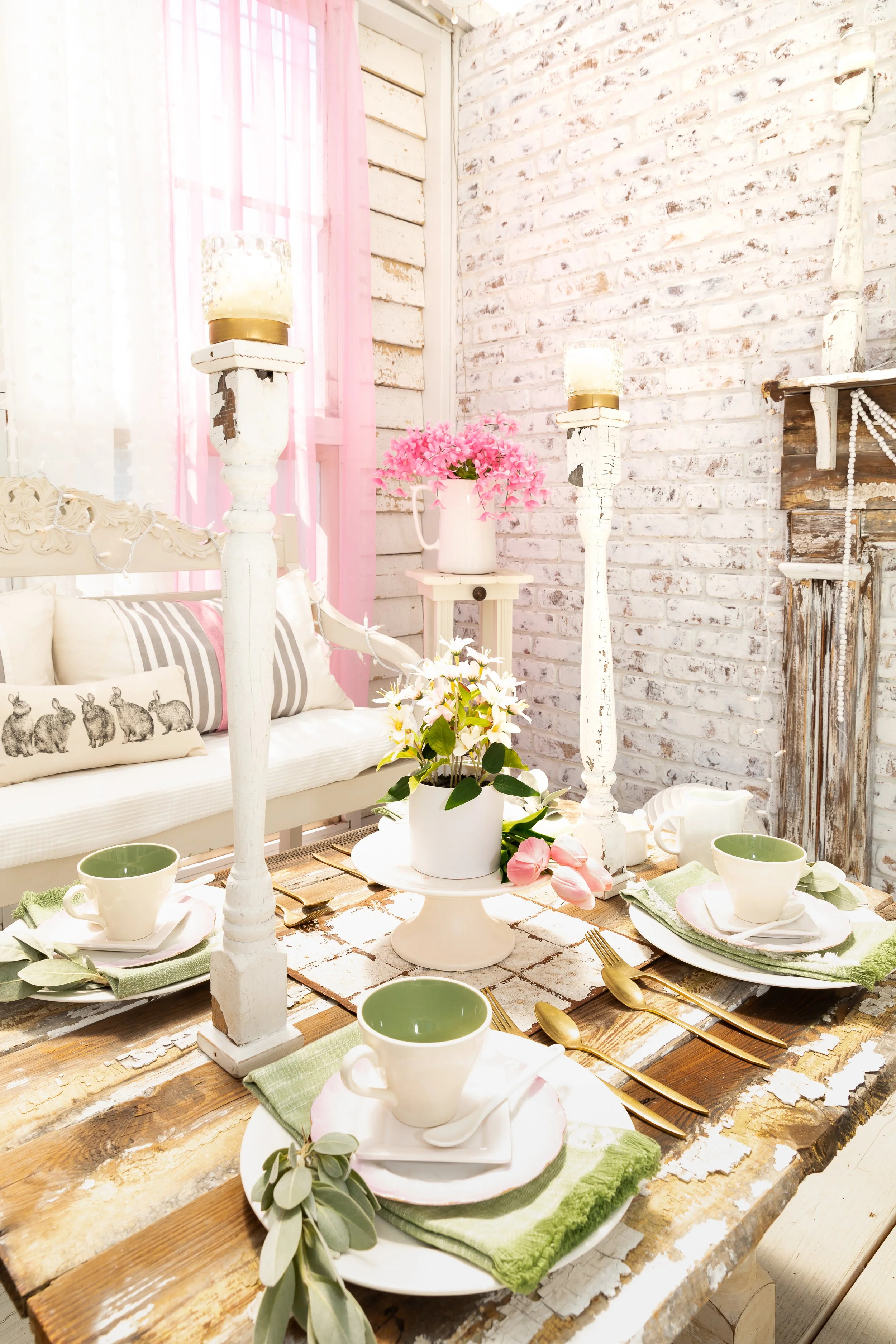 Rustic chic dining area with a distressed wood table set with white and green cups, gold cutlery, and decorative napkins. Candles on tall holders and pink flowers in a vase enhance the cozy ambiance, with a brick accent wall and a cushioned bench fea