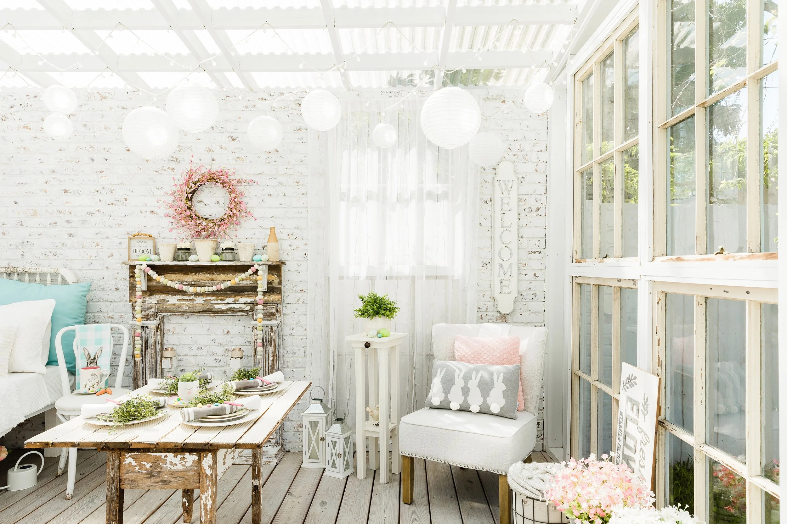 Brightly decorated studio with rustic white furniture, string lights, and paper lanterns. Features include a weathered table set with plates and greenery, a wreath and garland-adorned mantel, a chair with decorative pillows, and a "Welcome" sign.
