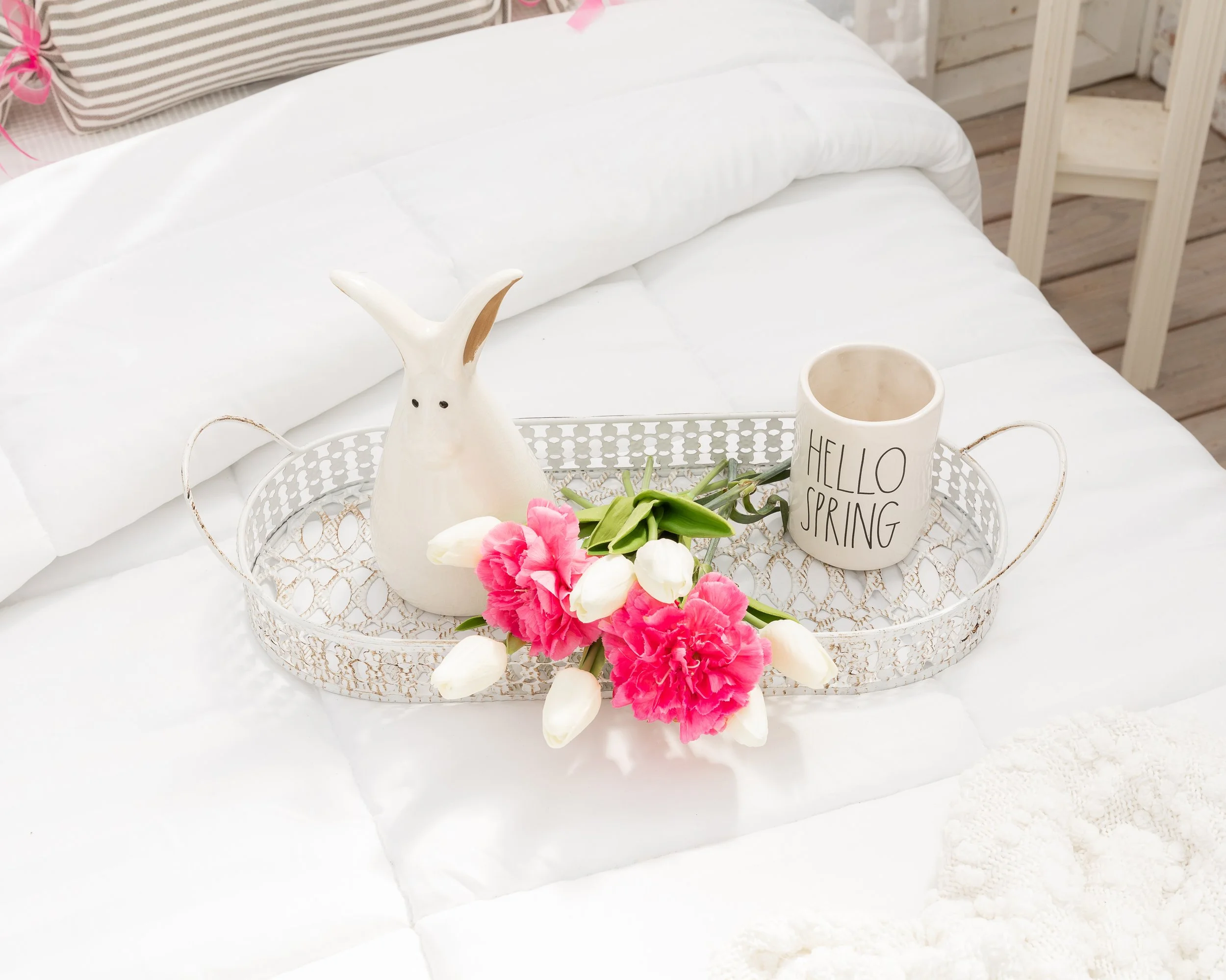 Decorative tray with a ceramic bunny, pink and white flowers, and a mug that says 'Hello Spring' on a white bed.