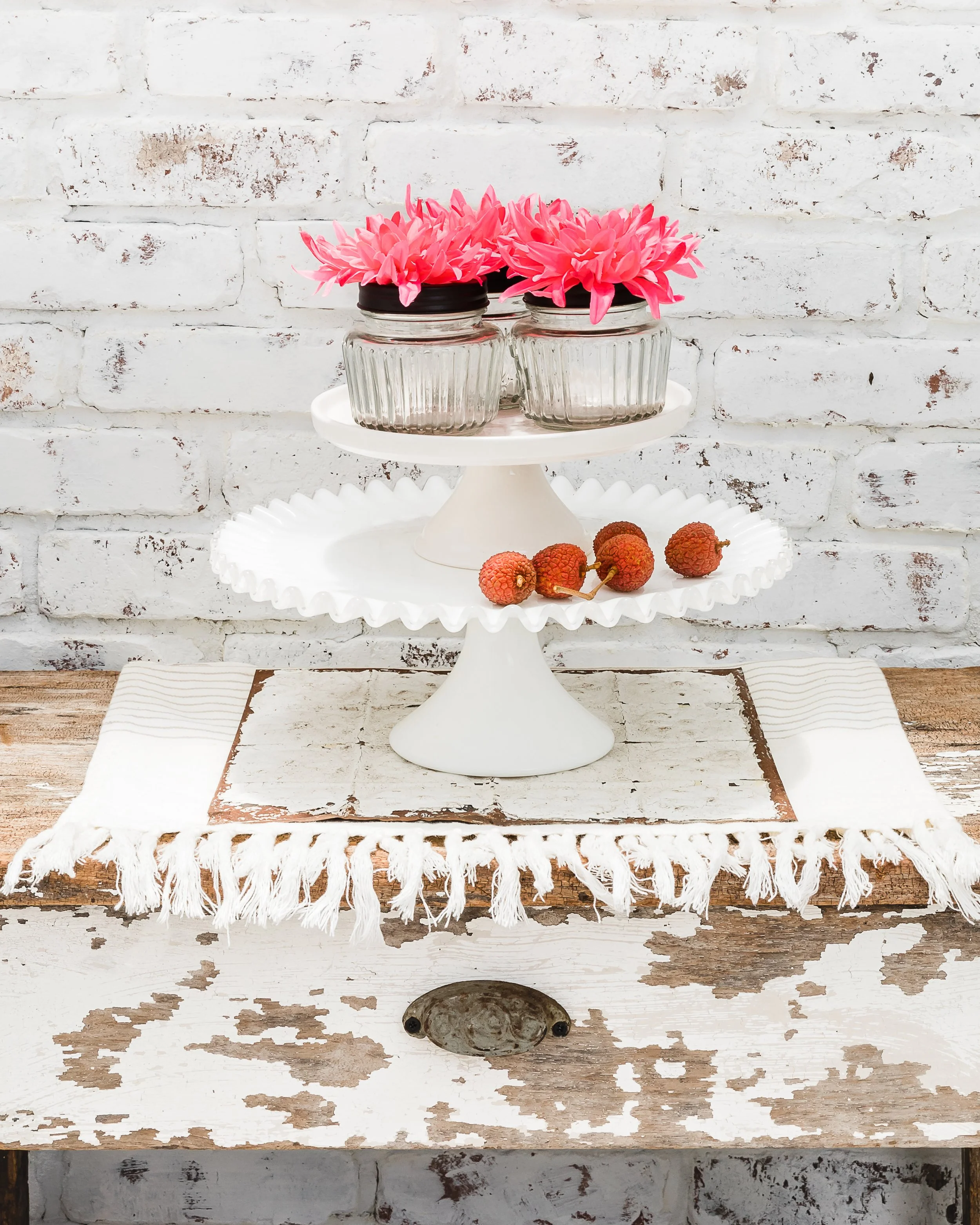 White tiered cake stand with pink flowers in glass jars on top and lychee fruit on the lower tier, placed on a rustic white wooden table against a white brick wall.