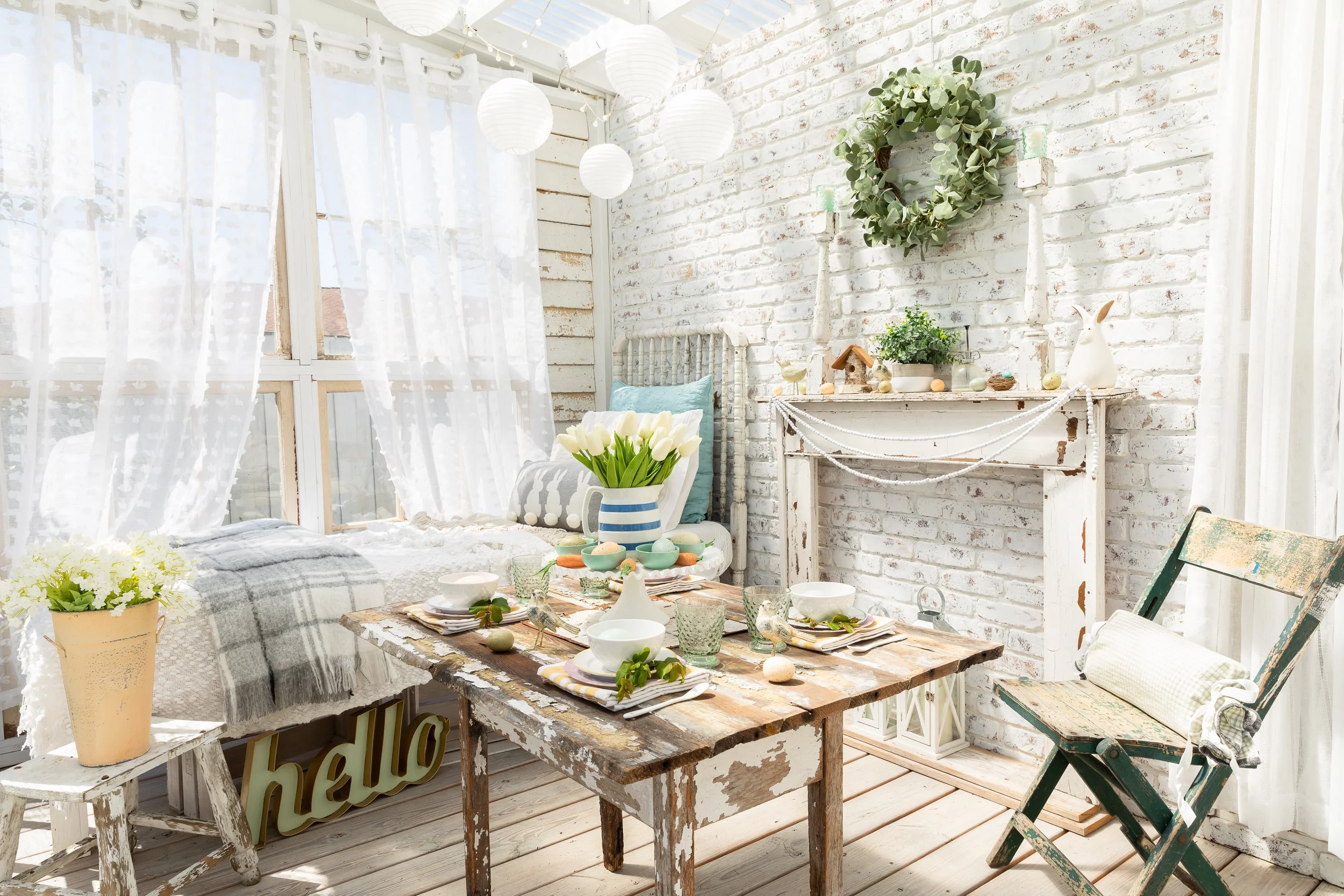 Bright and airy farmhouse-style room with whitewashed brick, rustic wooden table set for a meal, vase of tulips, distressed furniture, and decorative items including a wreath and paper lanterns.