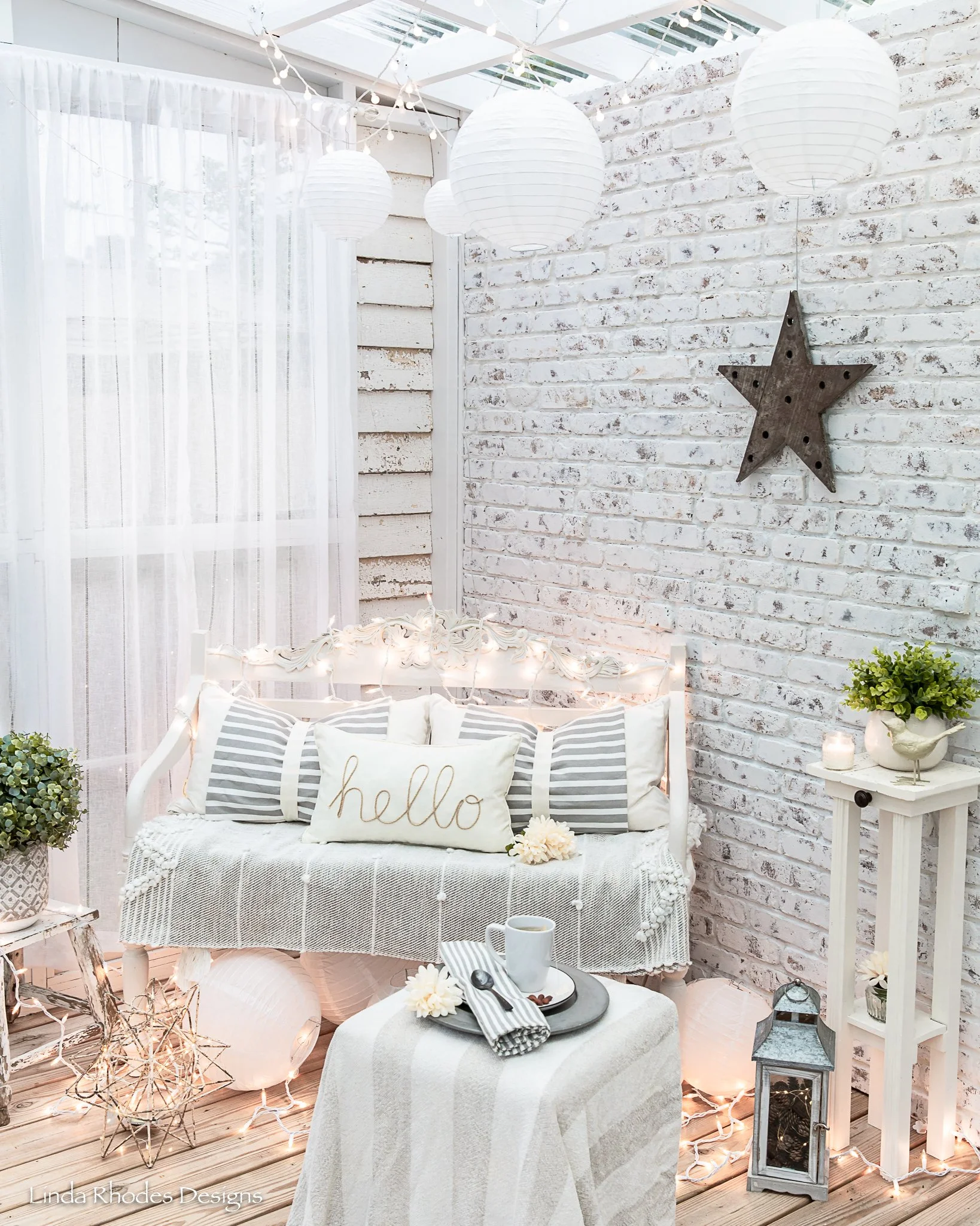A cozy, well-decorated sunroom with white brick walls, white lanterns hanging from the ceiling, and string lights. There's a white vintage-style bench with striped pillows and a cushion that says 'hello,' along with decorative flowers, a coffee cup, 