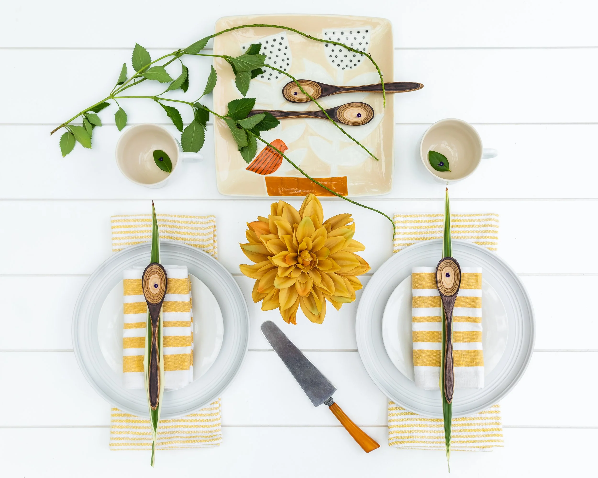 Table setting with two plates, yellow and white striped napkins, and wooden utensils decorated with leaves. A large yellow flower centerpiece and a flowering branch are on the table, with a cutting knife nearby. The table has a white surface.
