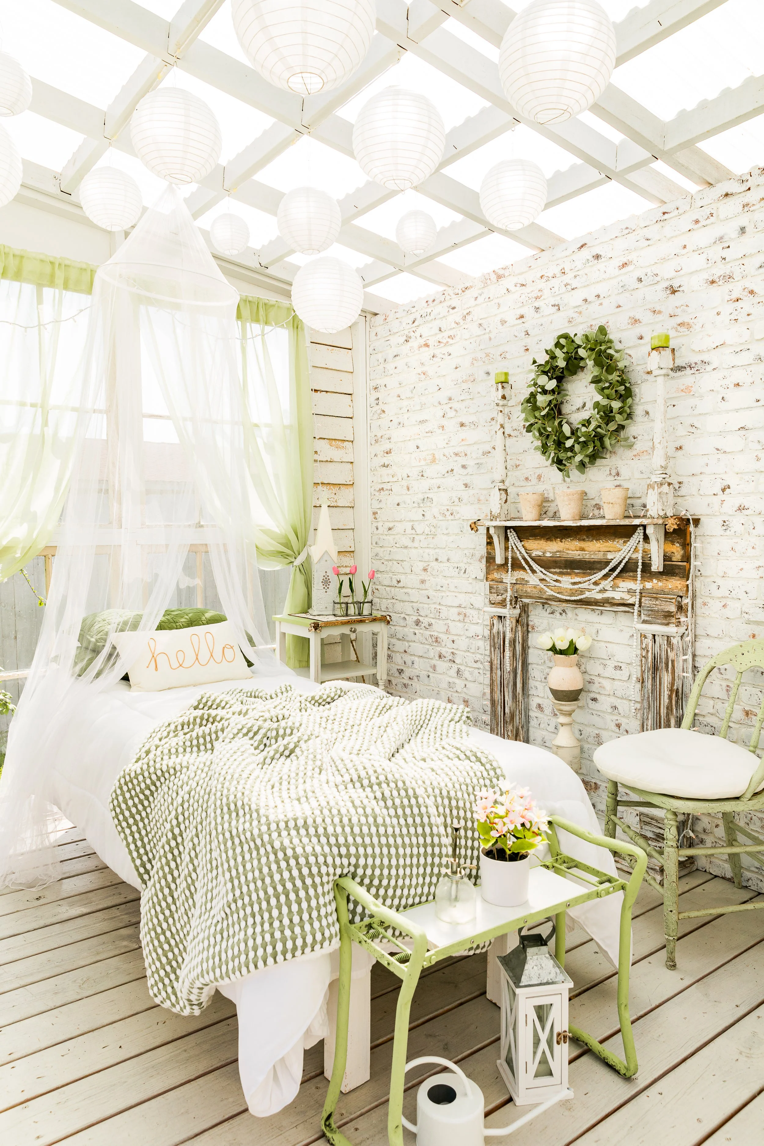 Bright and airy bedroom with a white canopy bed, green accents, and rustic decor. There are paper lanterns hanging from the ceiling, a wooden mantel with a wreath, and a green chair beside the bed. A small table holds a plant and there’s a watering c
