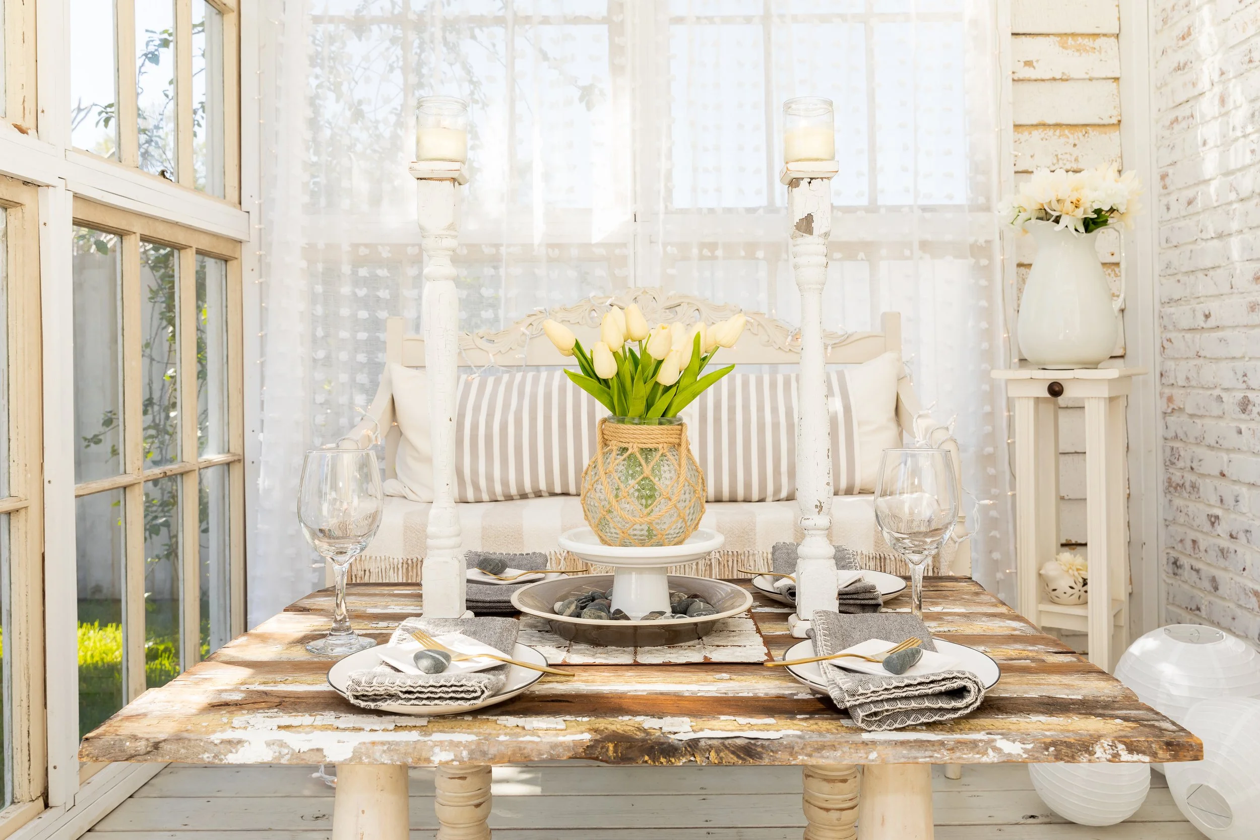 Rustic dining table setting with white candles, a vase of yellow tulips, glassware, and striped decor. Light, airy room with vintage furniture and window backdrop.