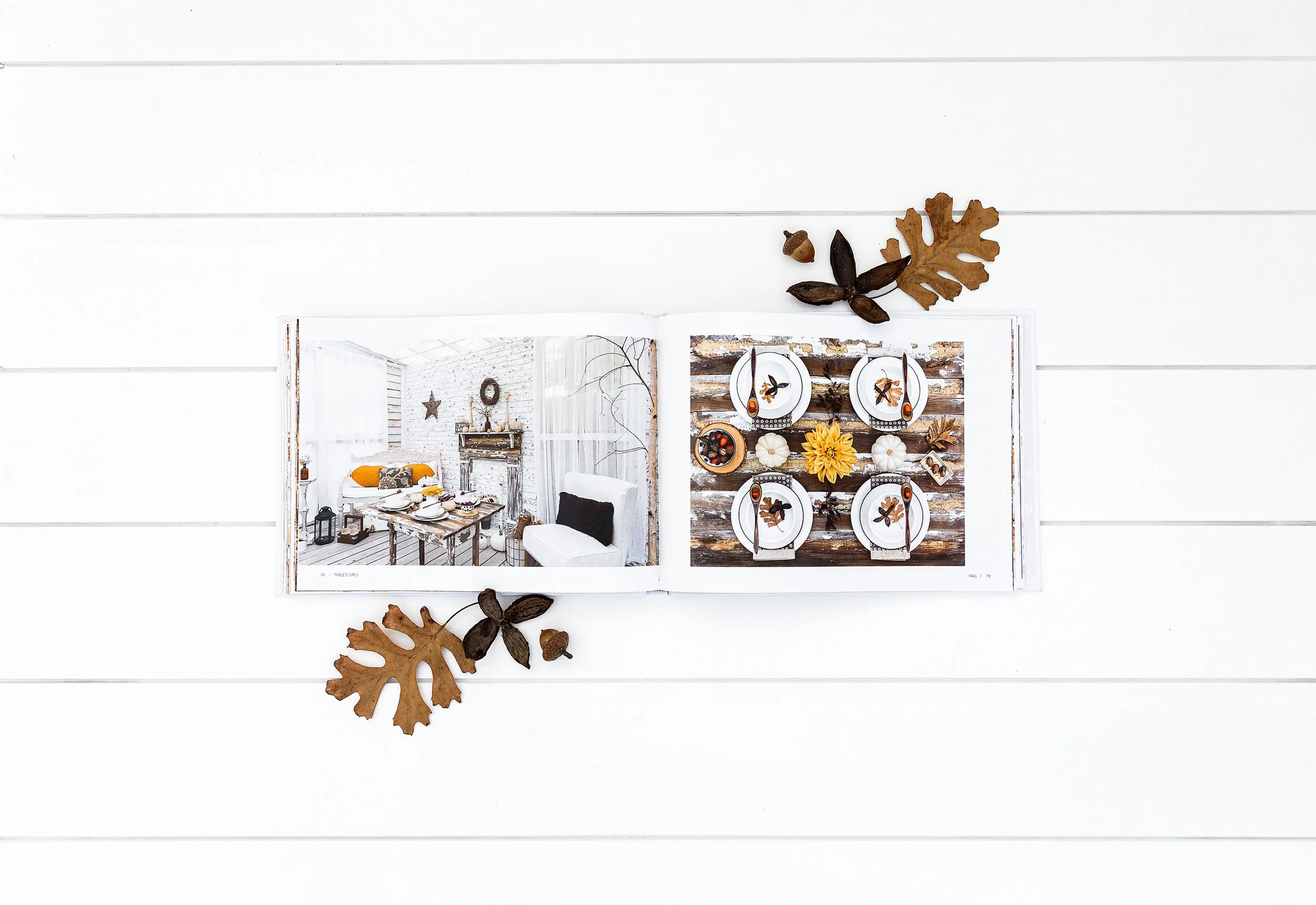 Open book displaying autumn-themed table setting and cozy living room, surrounded by dry autumn leaves on a white wooden surface.