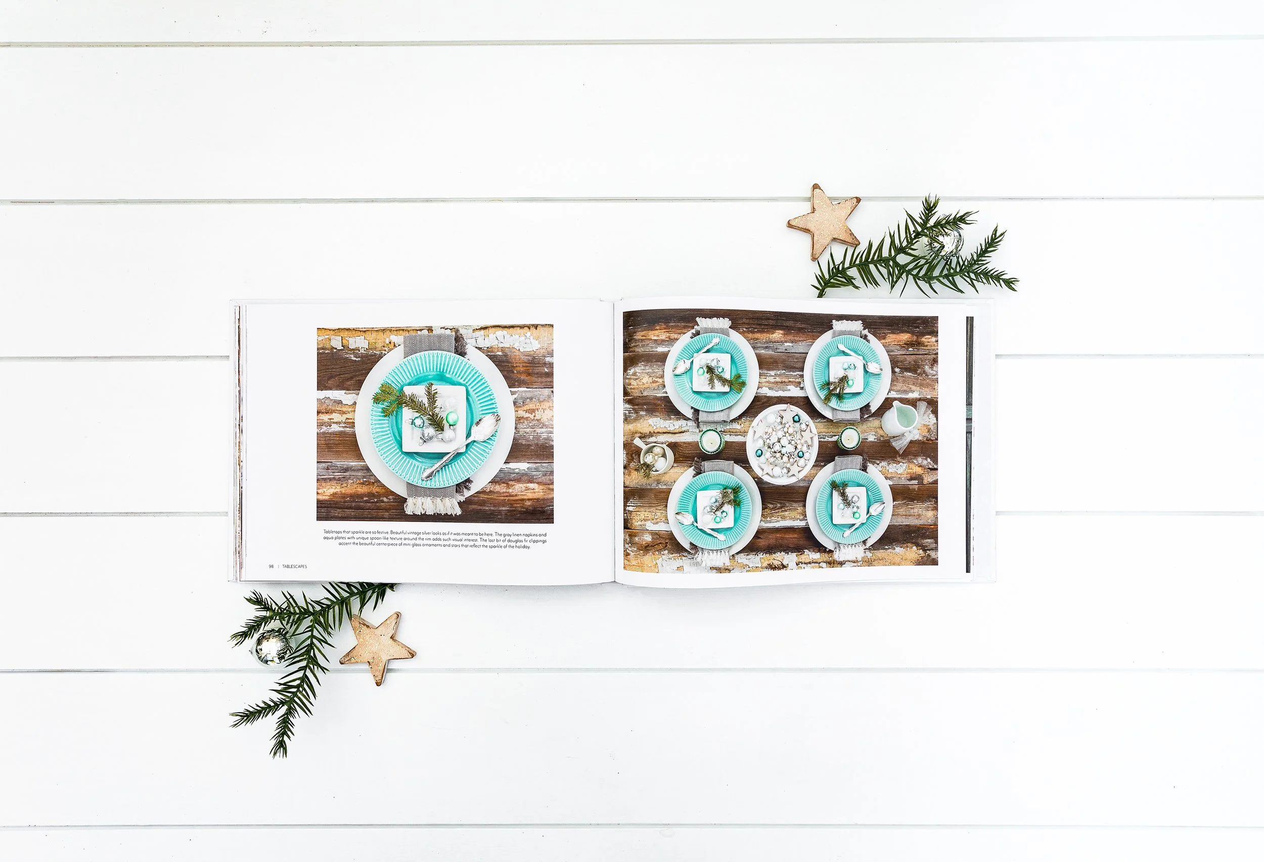 Open book on a white wooden surface displaying table setting images with teal plates, cutlery, and holiday decorations.
