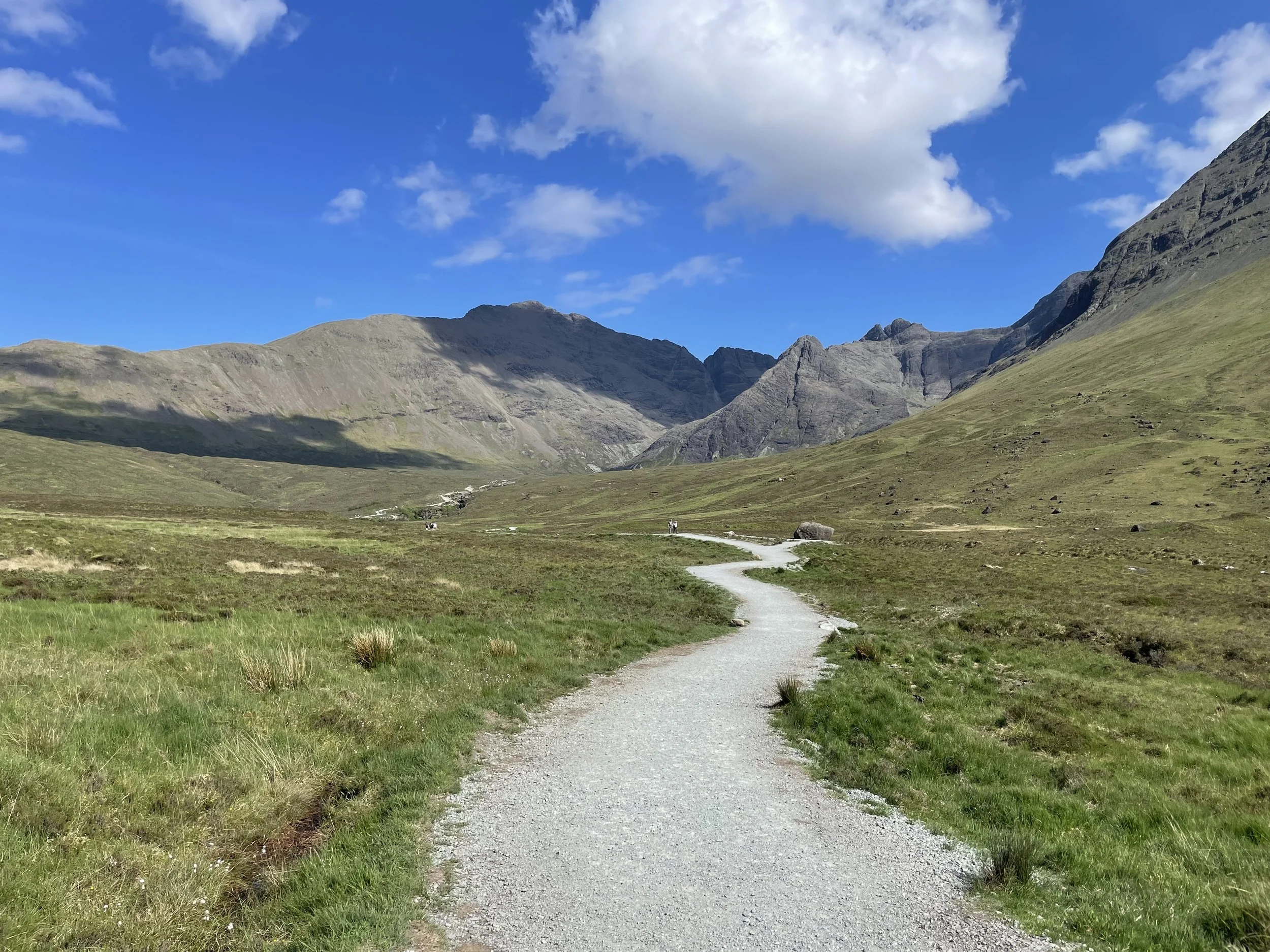 A winding gravel trail in a green valley with mountains in the background under a partly cloudy blue sky.
