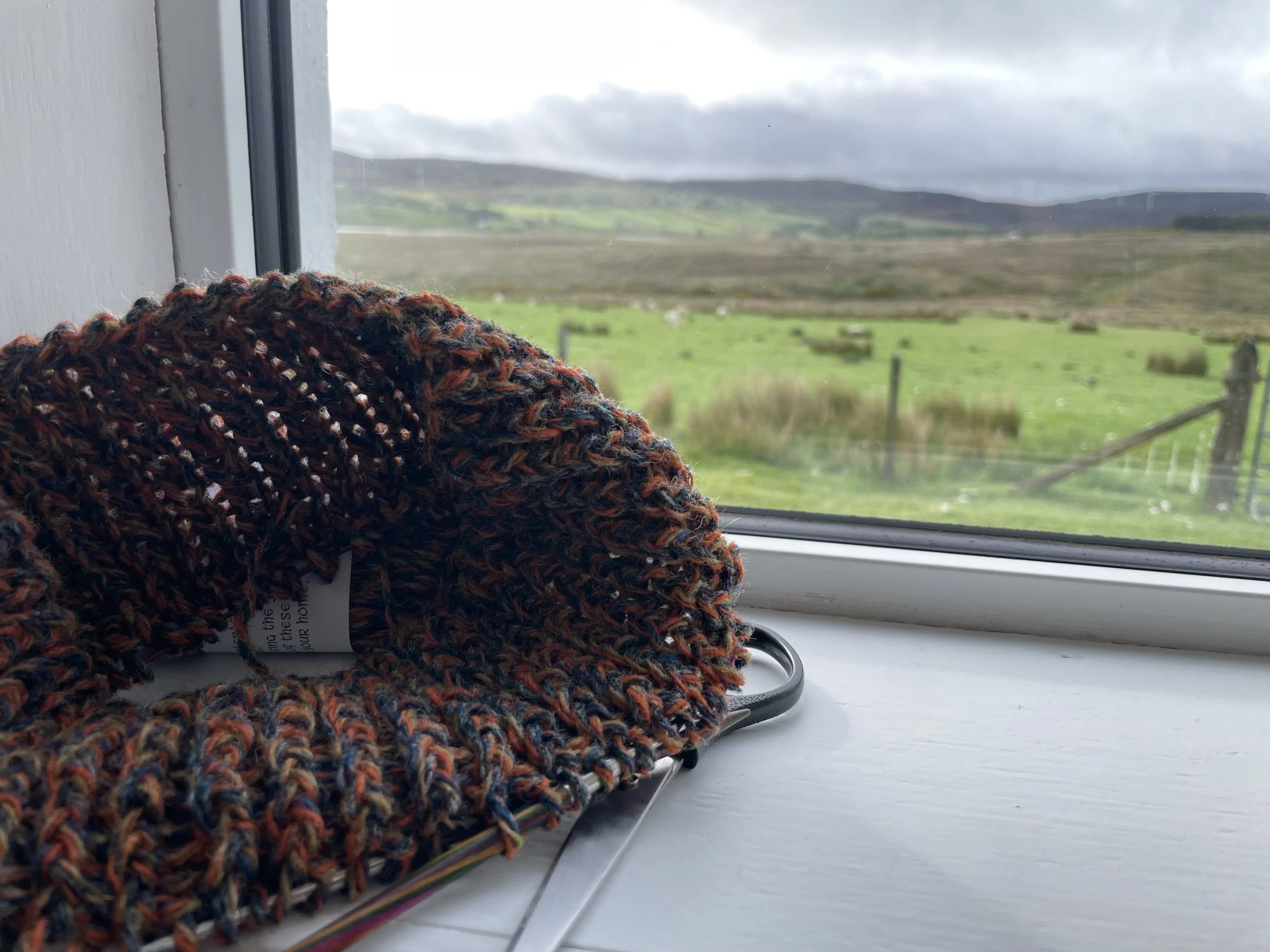 Colorful knitted hat resting on a windowsill with a view of Scotland's rolling green hills and cloudy sky outside.