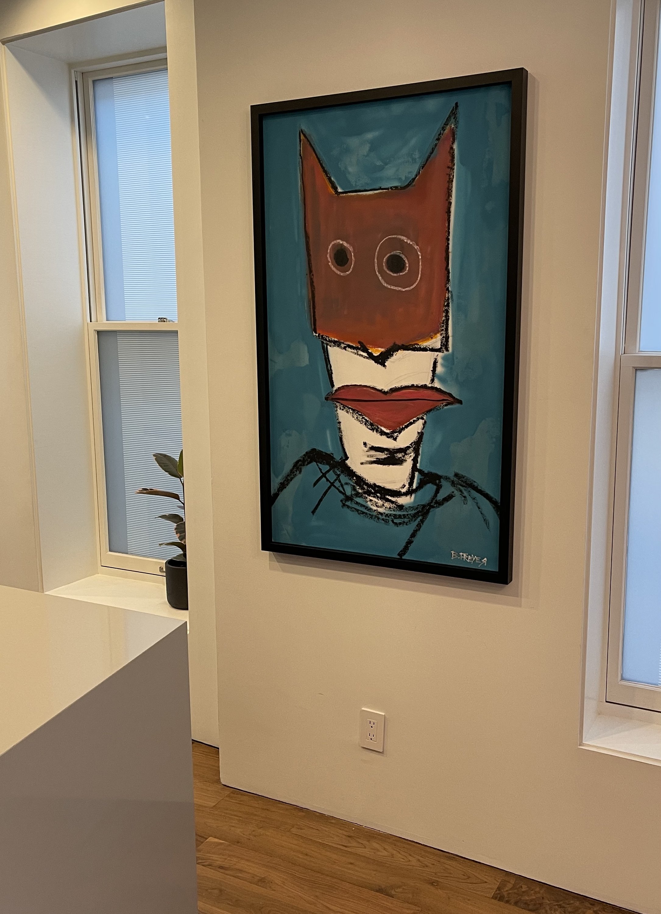 Framed abstract painting of a figure with a brown mask and big eyes on a teal background in a room with white walls and wooden floor.
