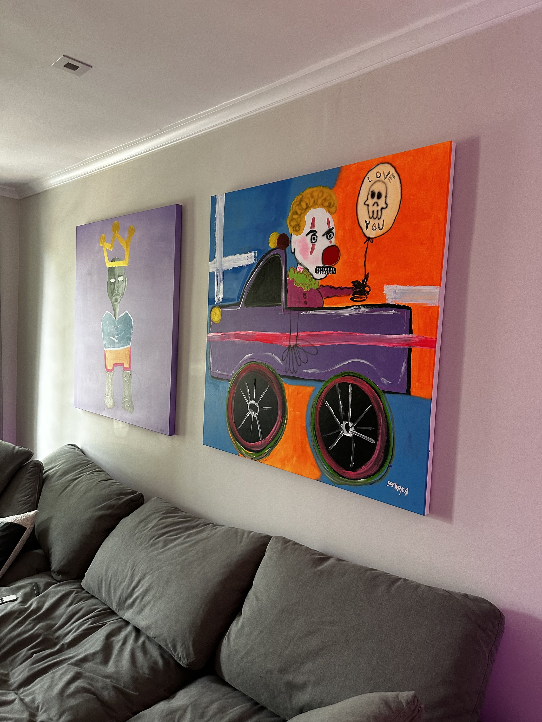 Two colorful abstract paintings on a wall above a gray couch. One painting depicts a character with a crown, while the other shows a clown-like figure holding a balloon with a skull and the words "Love You."