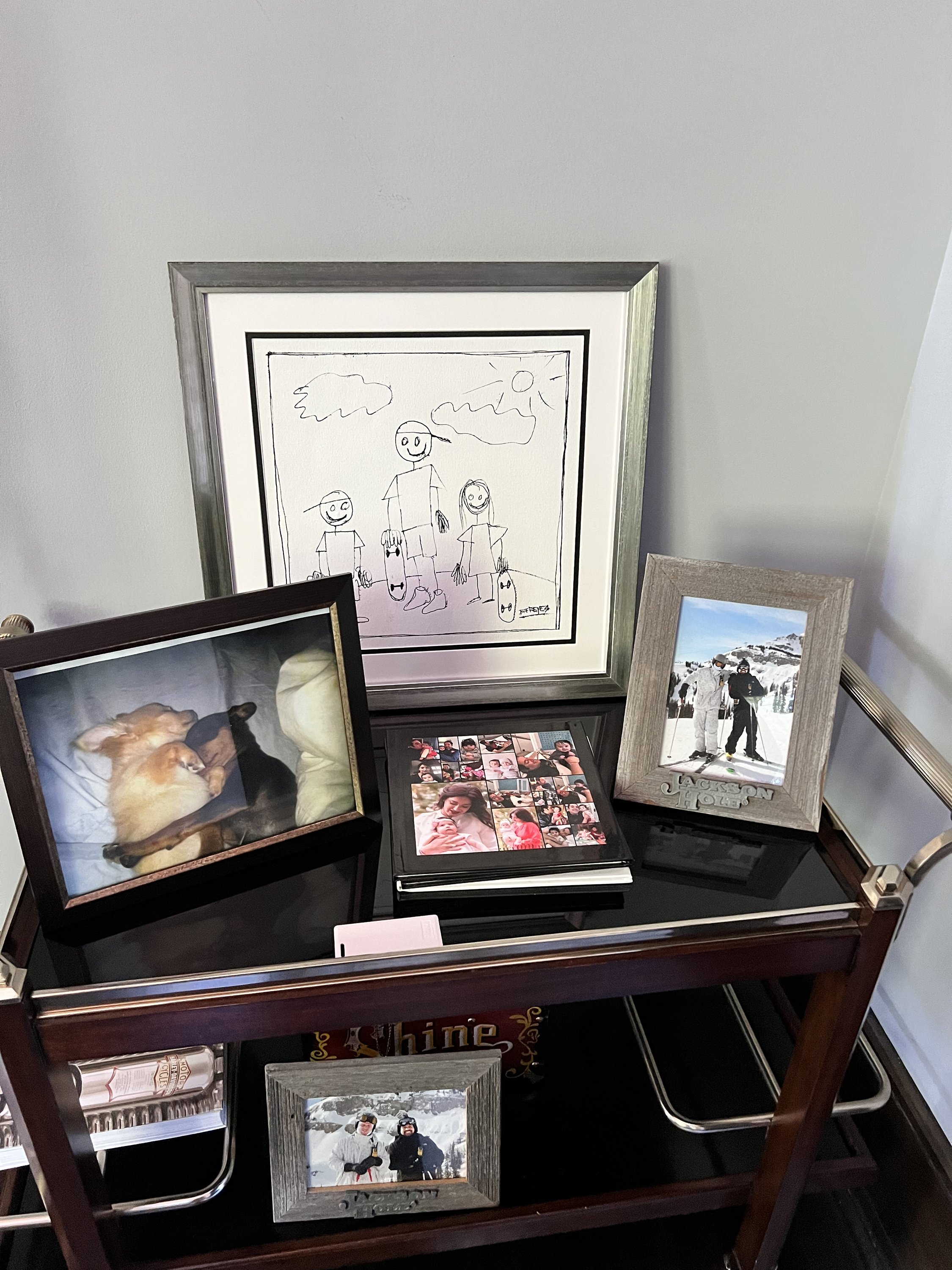 A small display table with a framed child's drawing, two framed photographs, a photo collage, and a home decor item labeled "Shine." The drawing shows three stick-figure people under a sun and clouds. The photos include one of two dogs and another of