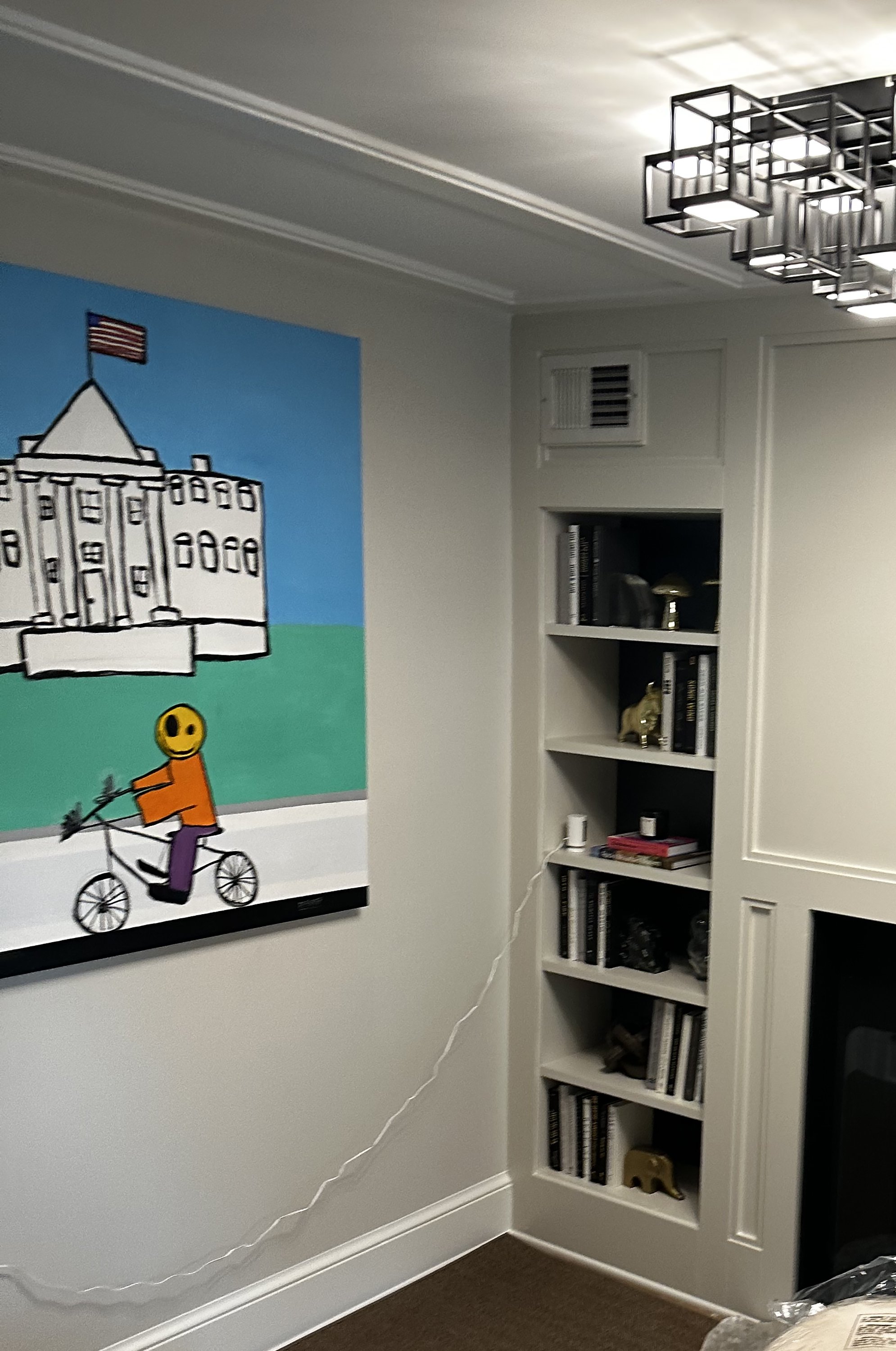 Interior room with a painting of a person on a bicycle in front of a building, shelves with books and decor, and a geometric ceiling light.