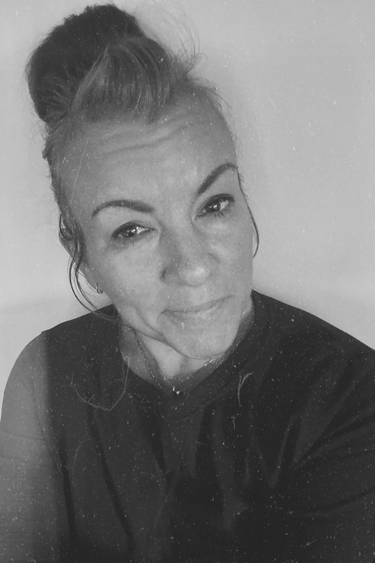 Black and white portrait of a person with a bun hairstyle, wearing a dark shirt, looking at the camera, with a textured vintage effect overlay.