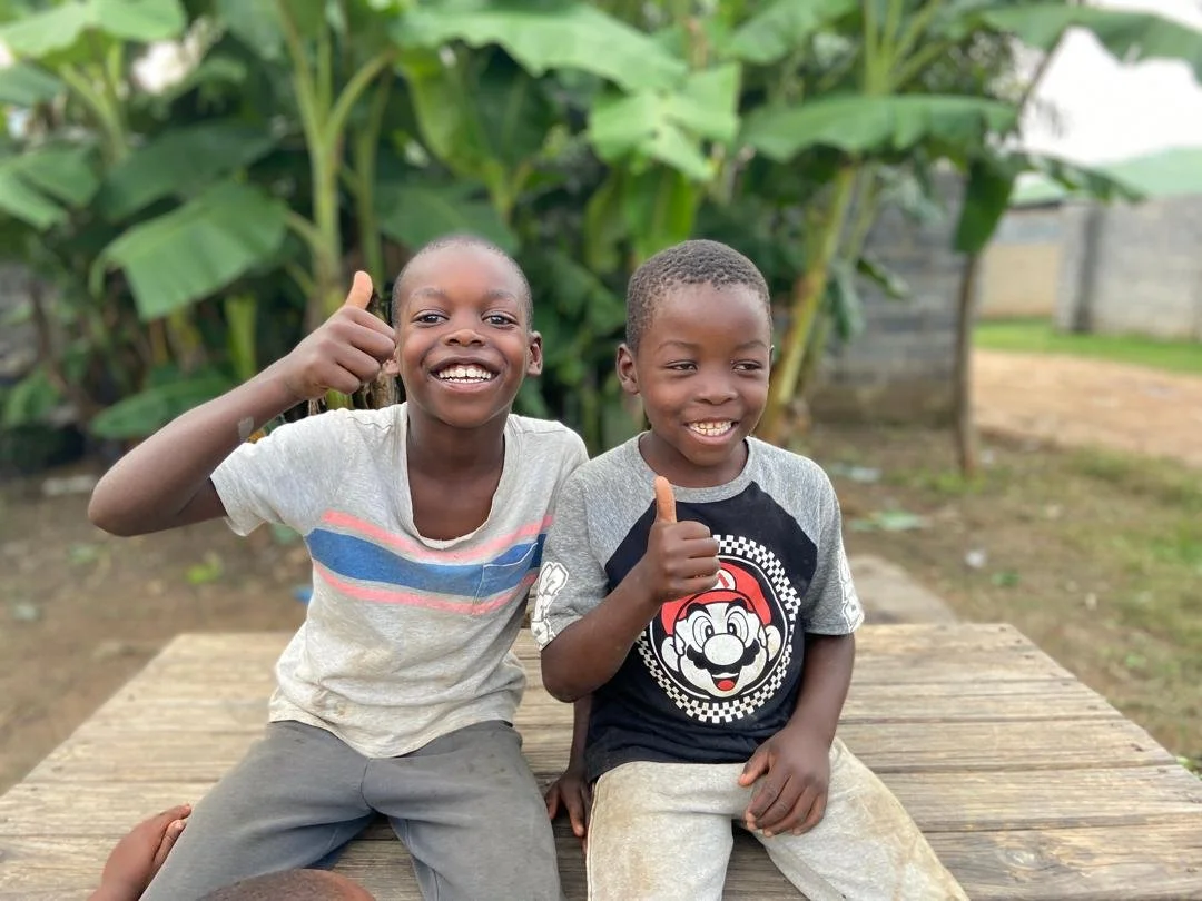 Two young boys sitting on a wooden platform outdoors, smiling, giving thumbs up, with greenery and banana plants in the background.
