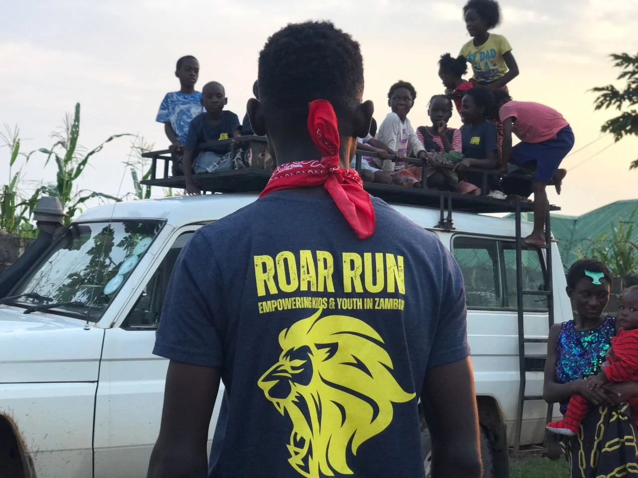 A young man with a red bandana on his neck stands in front of a white vehicle, with children on a platform above the vehicle. The young man wears a T-shirt with the text 'Roar Run Empowering Kids & Youth in Zambia' and a lion graphic. The children ar