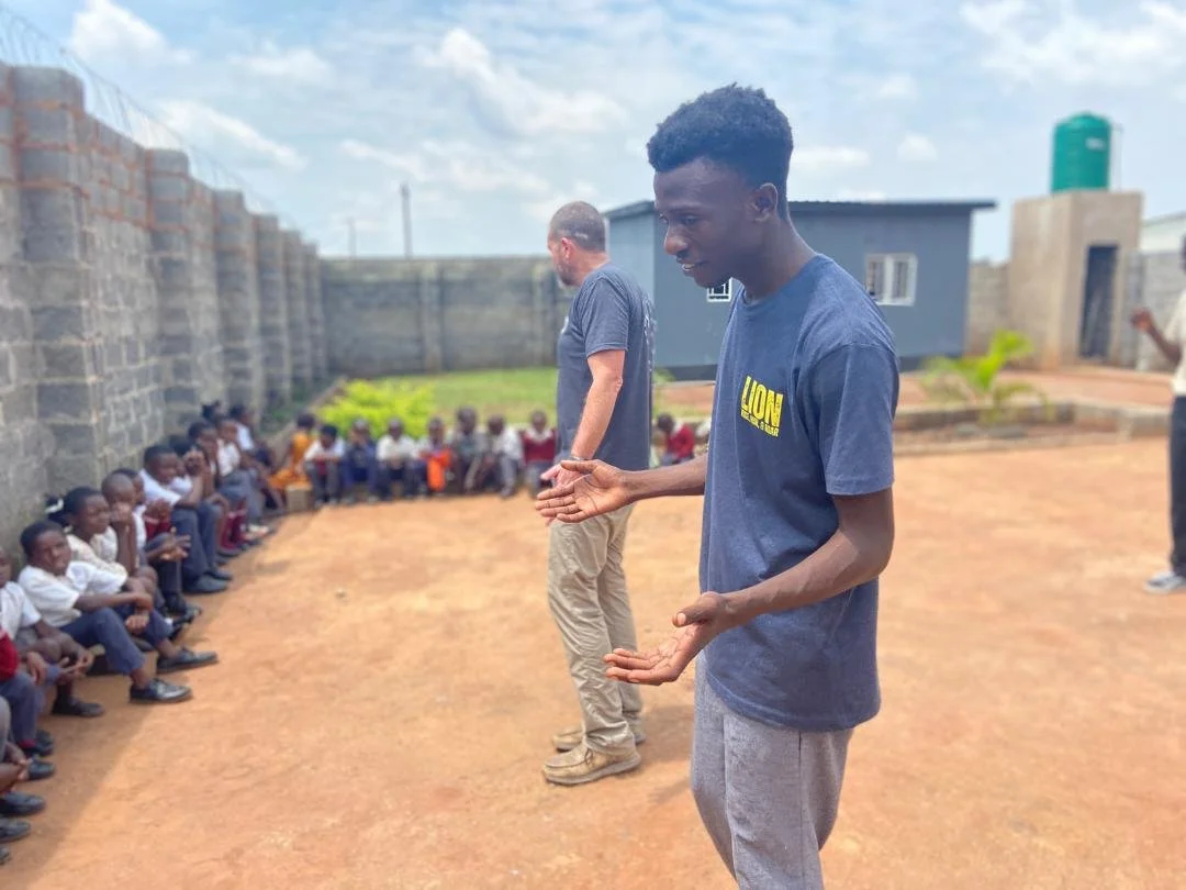 Two young men standing in front of a large group of children seated against a stone wall outdoors on a sunny day.