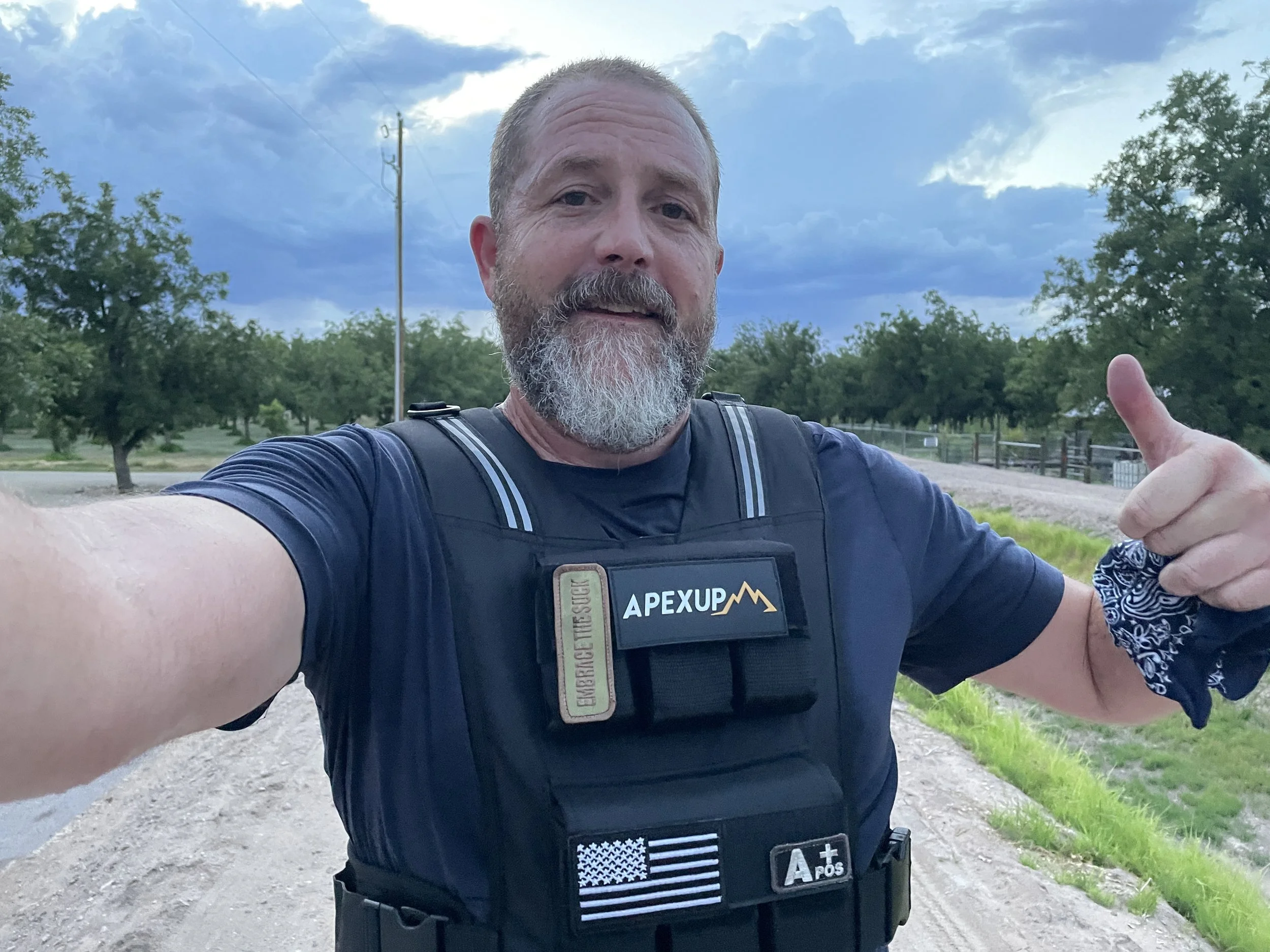 A man with a beard taking a selfie outdoors in a rural area with trees and cloudy sky, wearing a black tactical vest with patches, including one that says 'APEXUP' and another with an American flag.