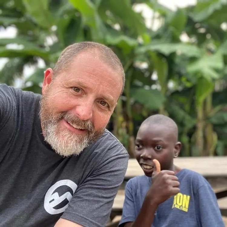A man with a beard and a gray T-shirt taking a selfie with a young boy giving a thumbs-up, with greenery in the background.