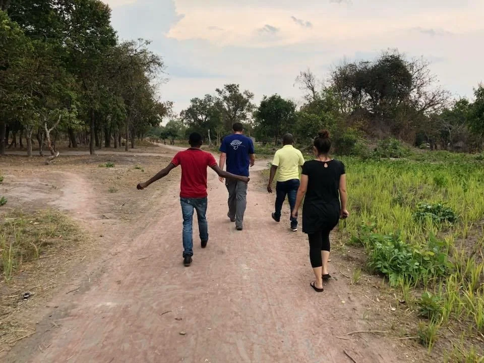Five people walking on a dirt path in a rural area with trees and grassy fields on either side.