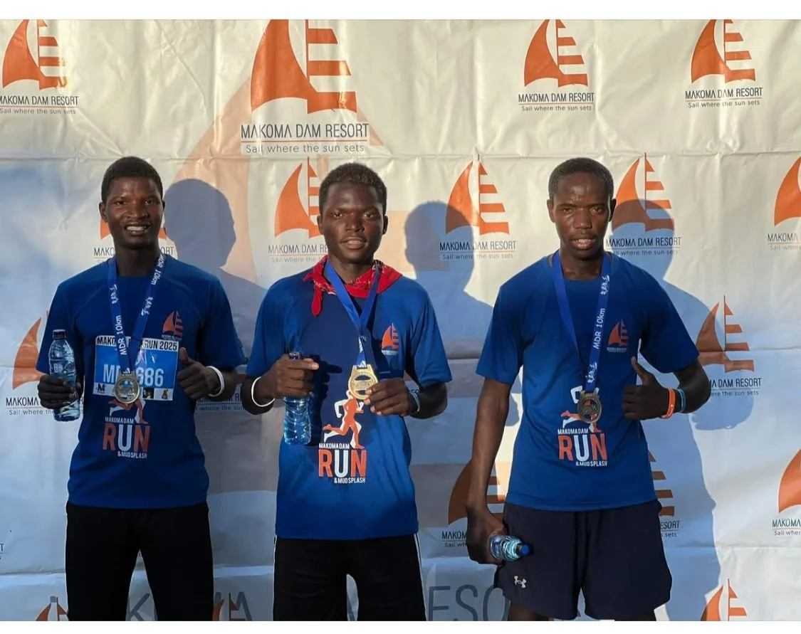 Three male runners on a podium holding water bottles and medals, wearing blue race shirts with the event logo, in front of a backdrop with the 'Makoma Dam Resort' logo.