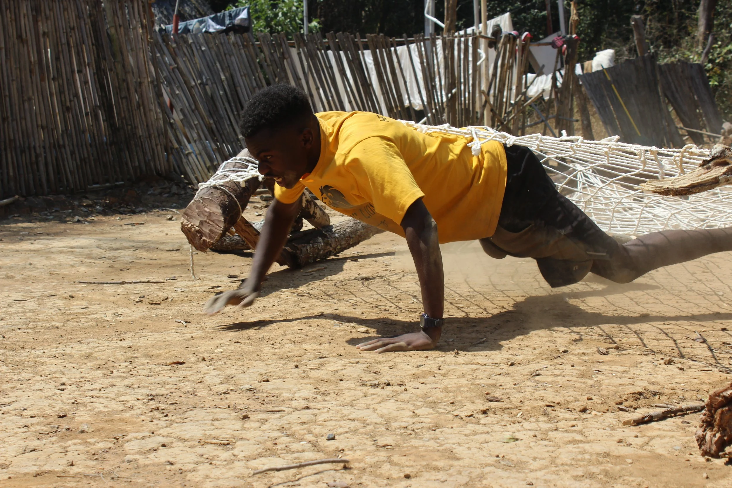 Young man in a yellow T-shirt performing a push-up on dirt ground with a wooden log and a fence in the background.