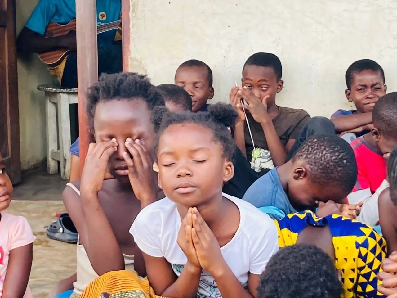 A group of children sitting on the ground with their eyes closed, some with hands in prayer or meditative poses, in an indoor setting with a plain wall in the background.