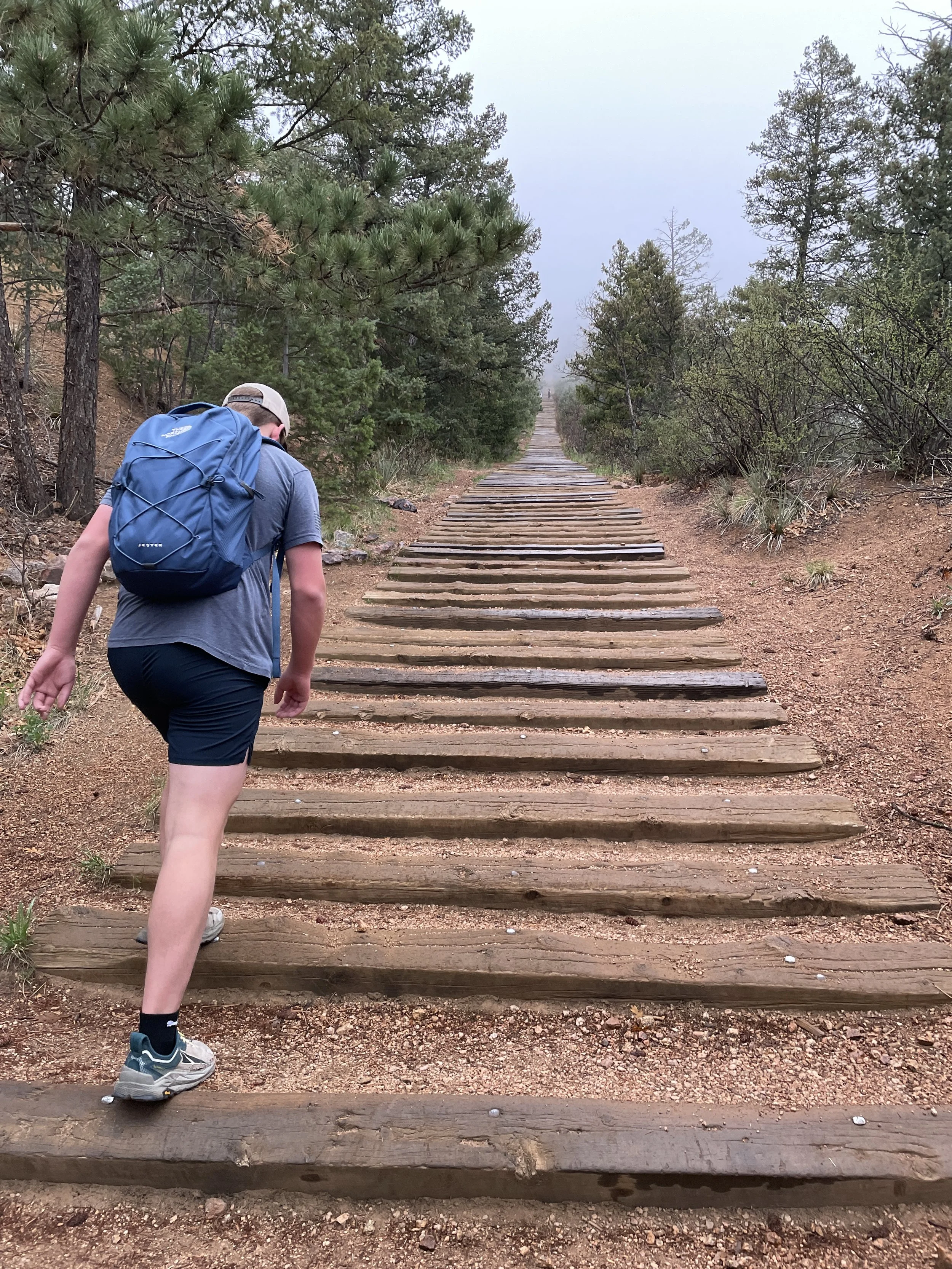 Hiker with a blue backpack walking on a wooden trail through a forest with fog.