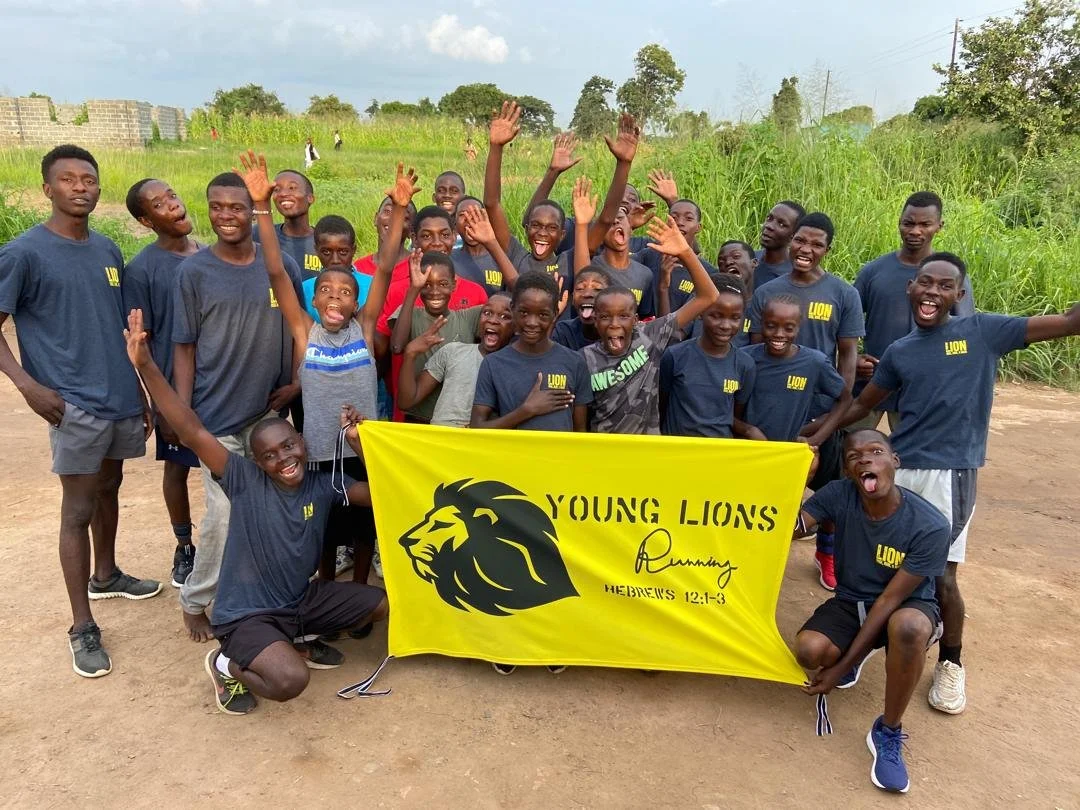 A group of young boys and teens posing outdoors on a dirt field with a green grassy background, holding a yellow flag with a lion logo and the words "Young Lions" and a Bible verse reference."